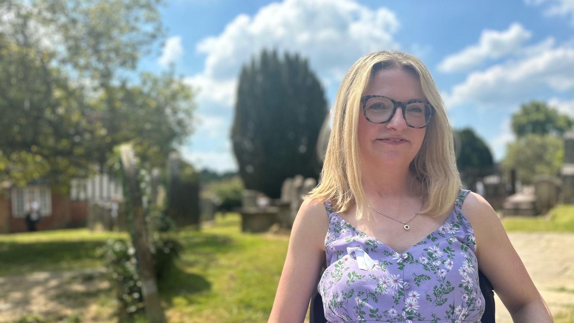 A woman in a purple green and white dress in a church yard. The woman is blonde, has brown glasses and is looking at the camera.