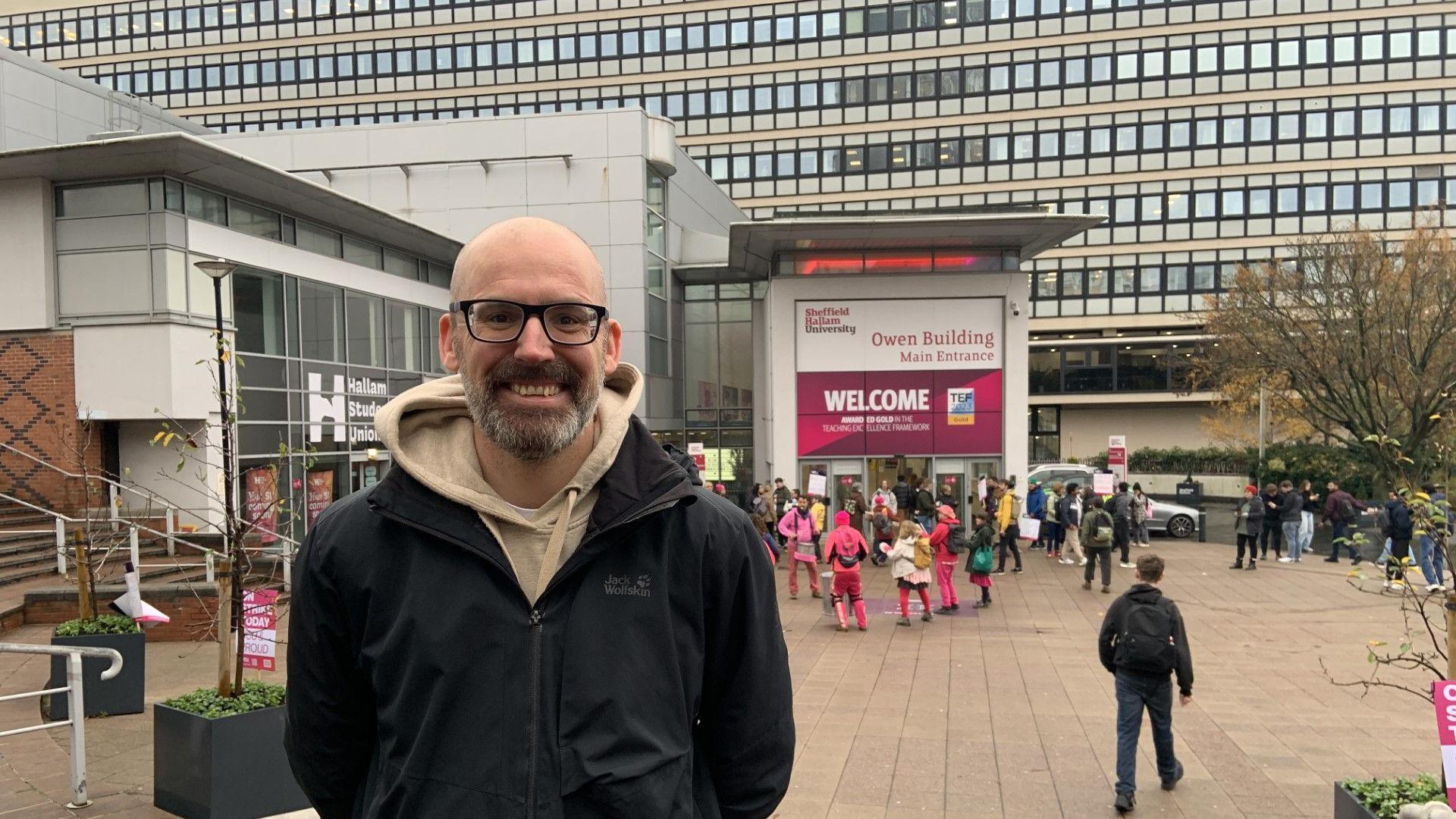 A man with a bald head, a beard and glasses is wearing a beige coloured hoodie and a black coat. He is standing outside Sheffield Hallam University, in the background is a picket line and a musical band with people dressed in pink