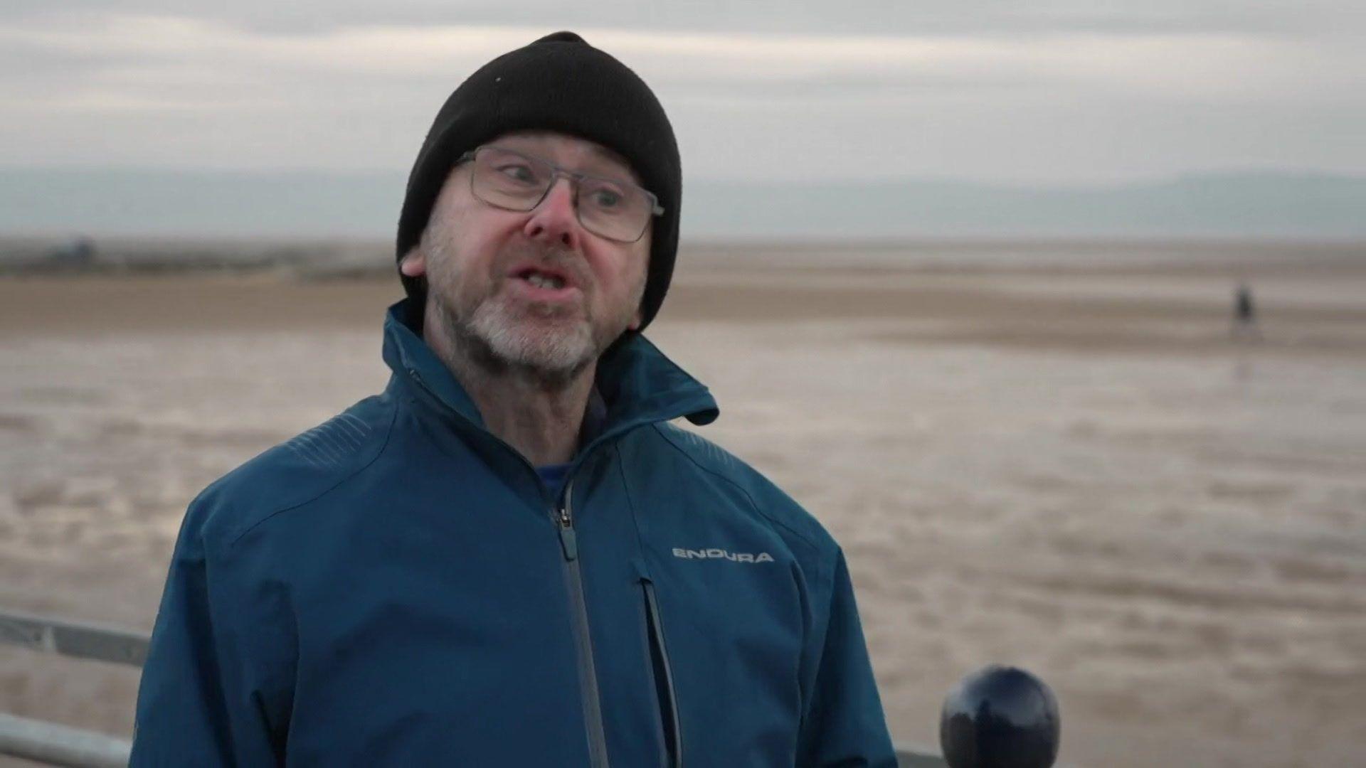 Wirral Green councillor Pat Cleary stands on the promenade at Leasowe. He is wearing a blue jacket and a blue woolly hat. 