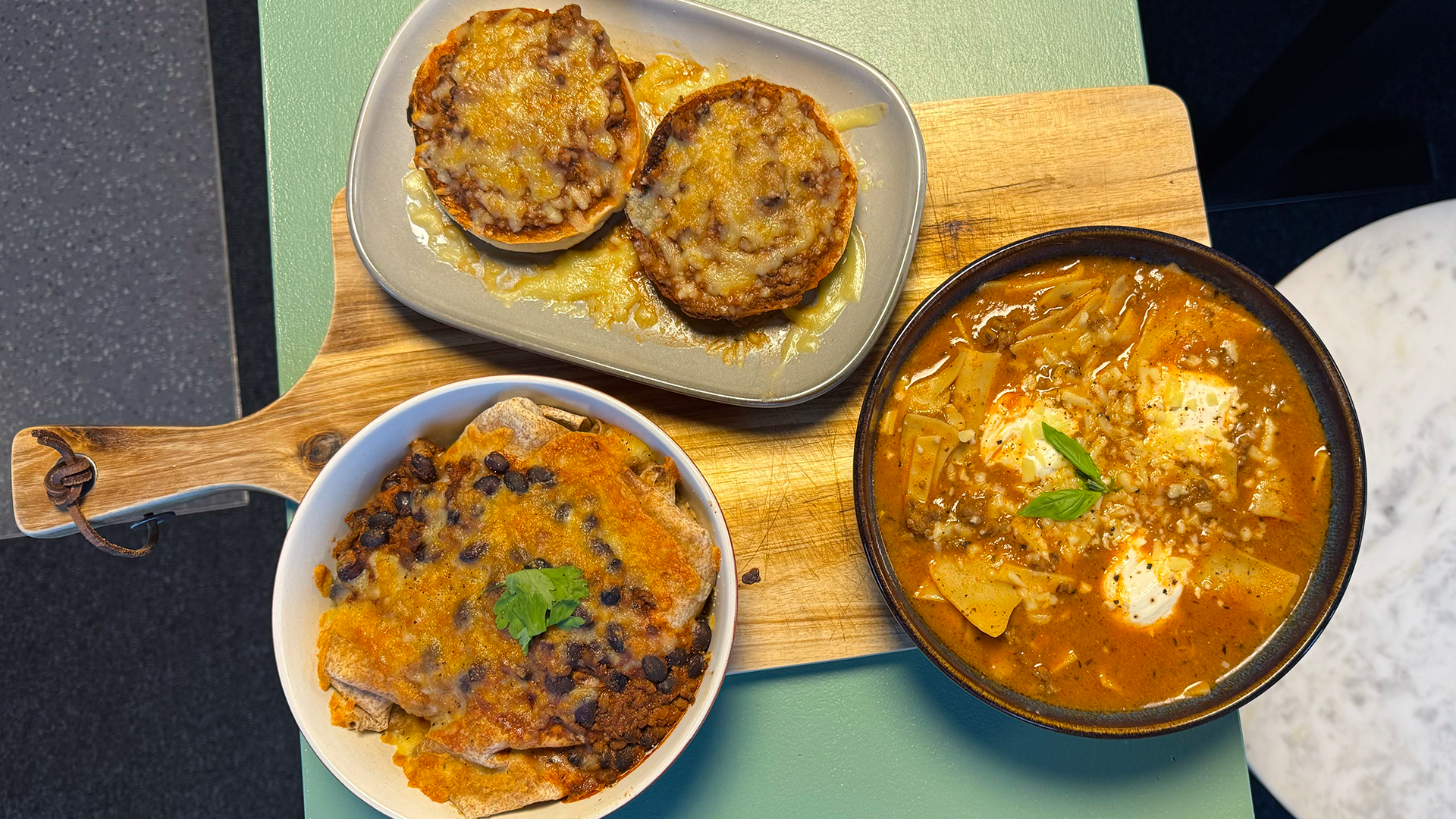 Briony's three bolognese meals on a wooden board. A dish of bolognese enchiladas, a bowl of lasagna soup and two cheesy pizza bagels on a ceramic plate.
