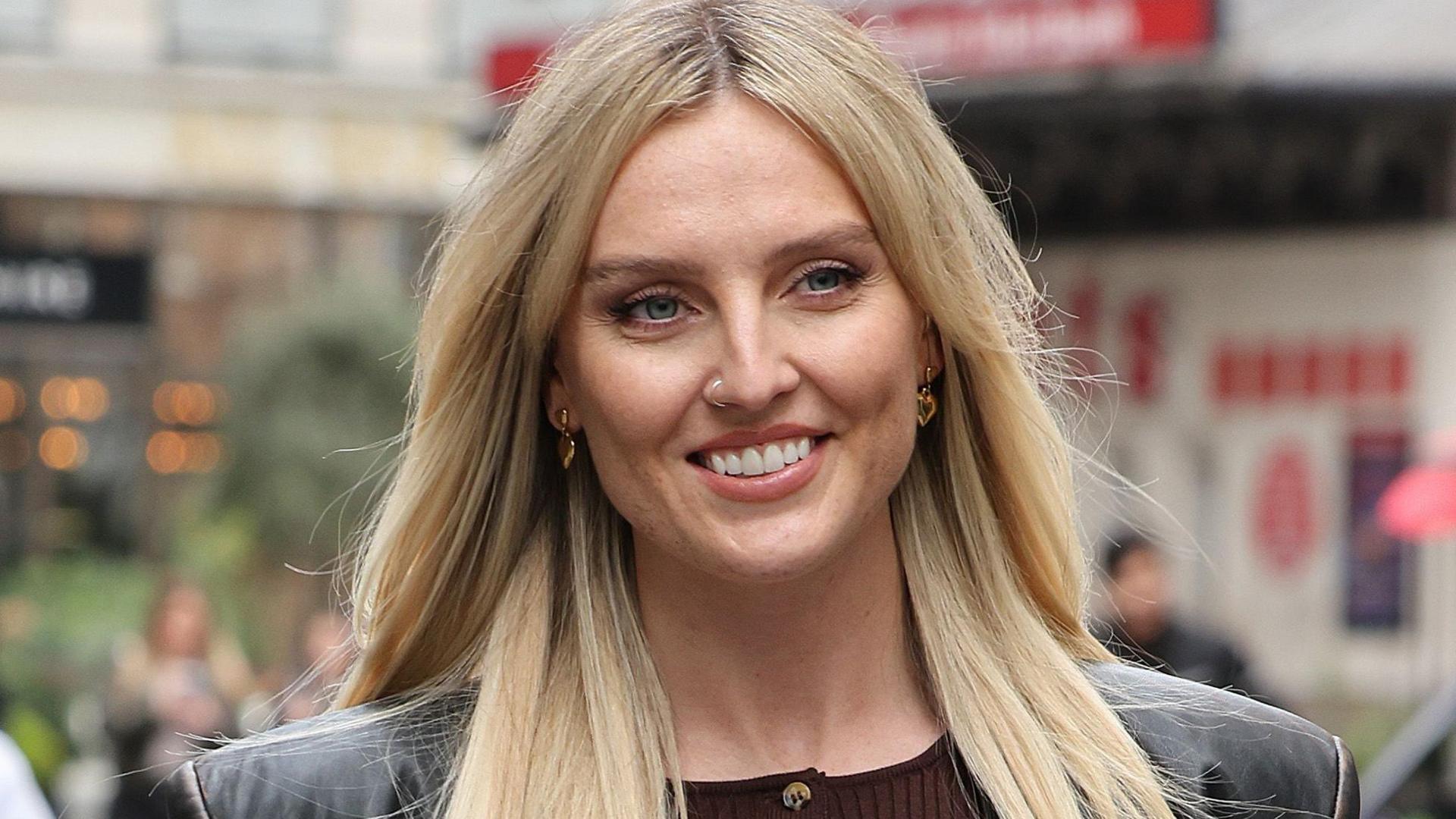 Perrie Edwards pictured out walking in London. Perrie has long blonde hair worn loose and blue eyes. She wears a black leather jacket over a brown top. 