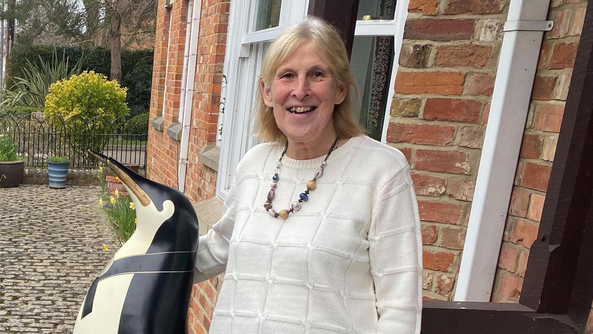 A woman wearing a white jumper and a necklace standing outside her house with her arm around a large penguin decoration made of glass fibre.