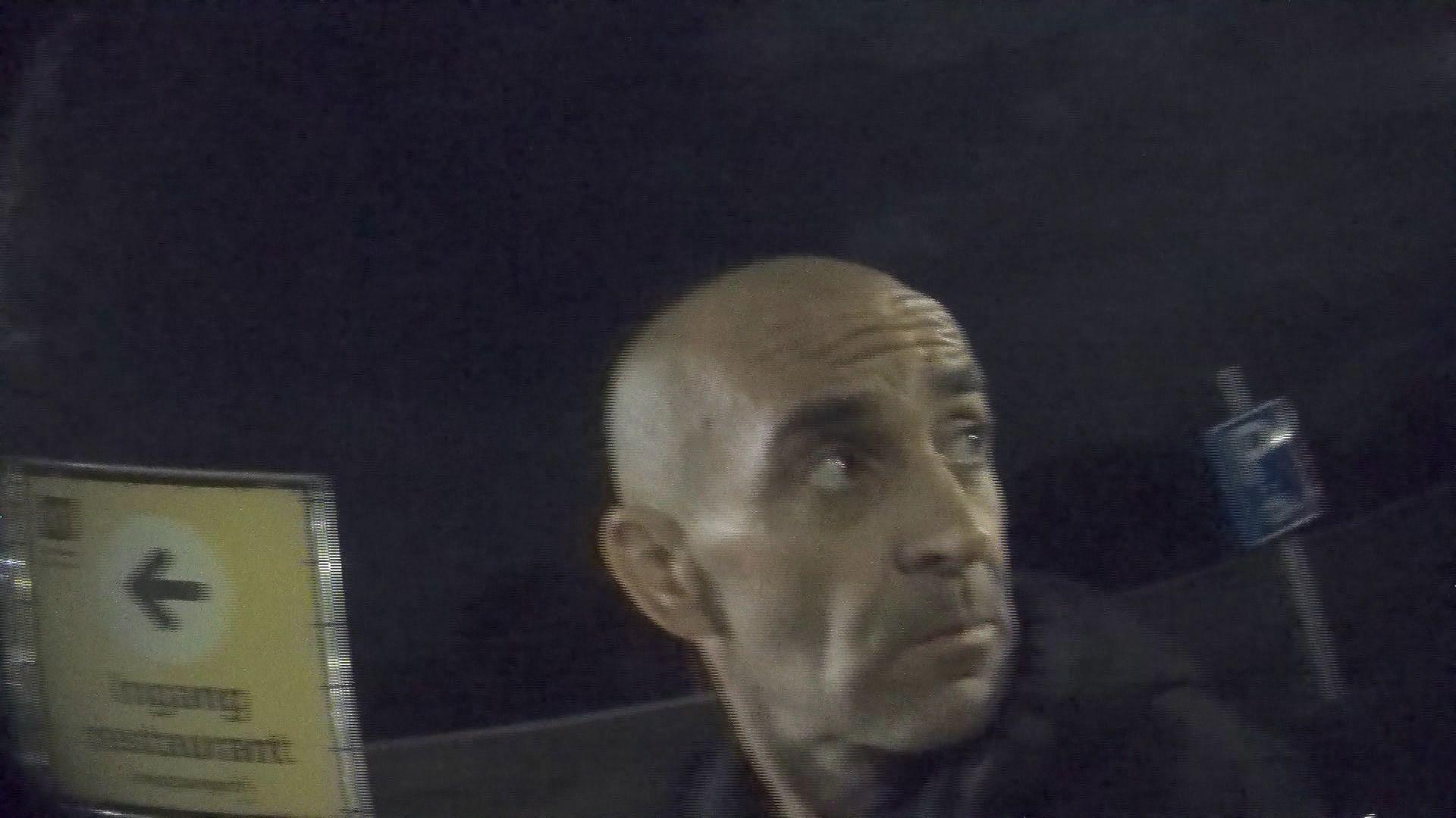 A man is looking to the right of the camera in a grainy image taken from an undercover camera. He is bald with large black eyebrows. Behind the man is a yellow sign for a restaurant and a blue parking sign, indicating he is in a car park. 
