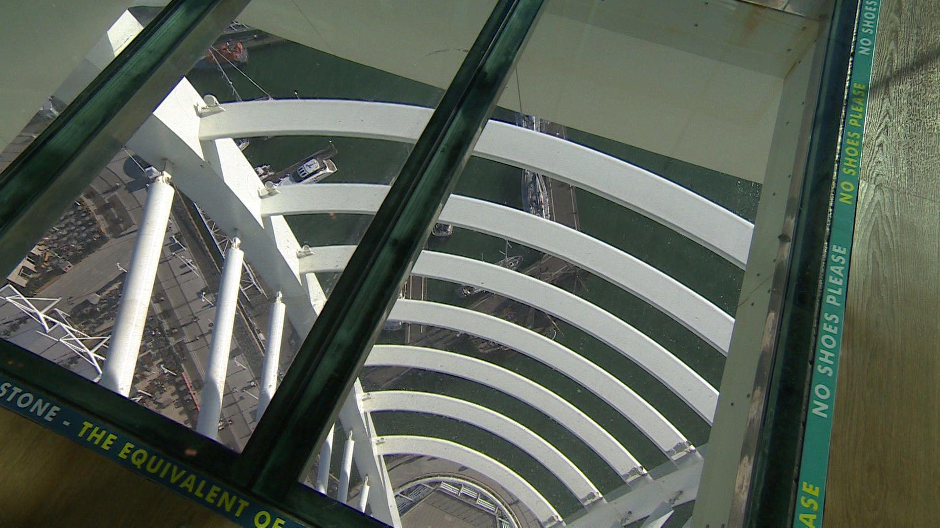 View through the glass floor of the Spinnaker Tower, showing the ground and water far below, with "NO SHOES PLEASE" text around the edges.