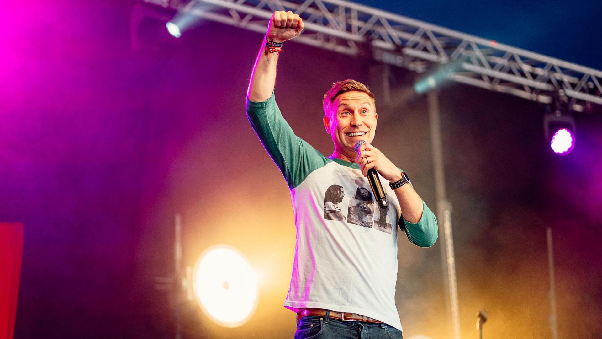 Russell Howard raises a triumphant fist on stage during his set. He's wearing a long sleeved top and jeans.