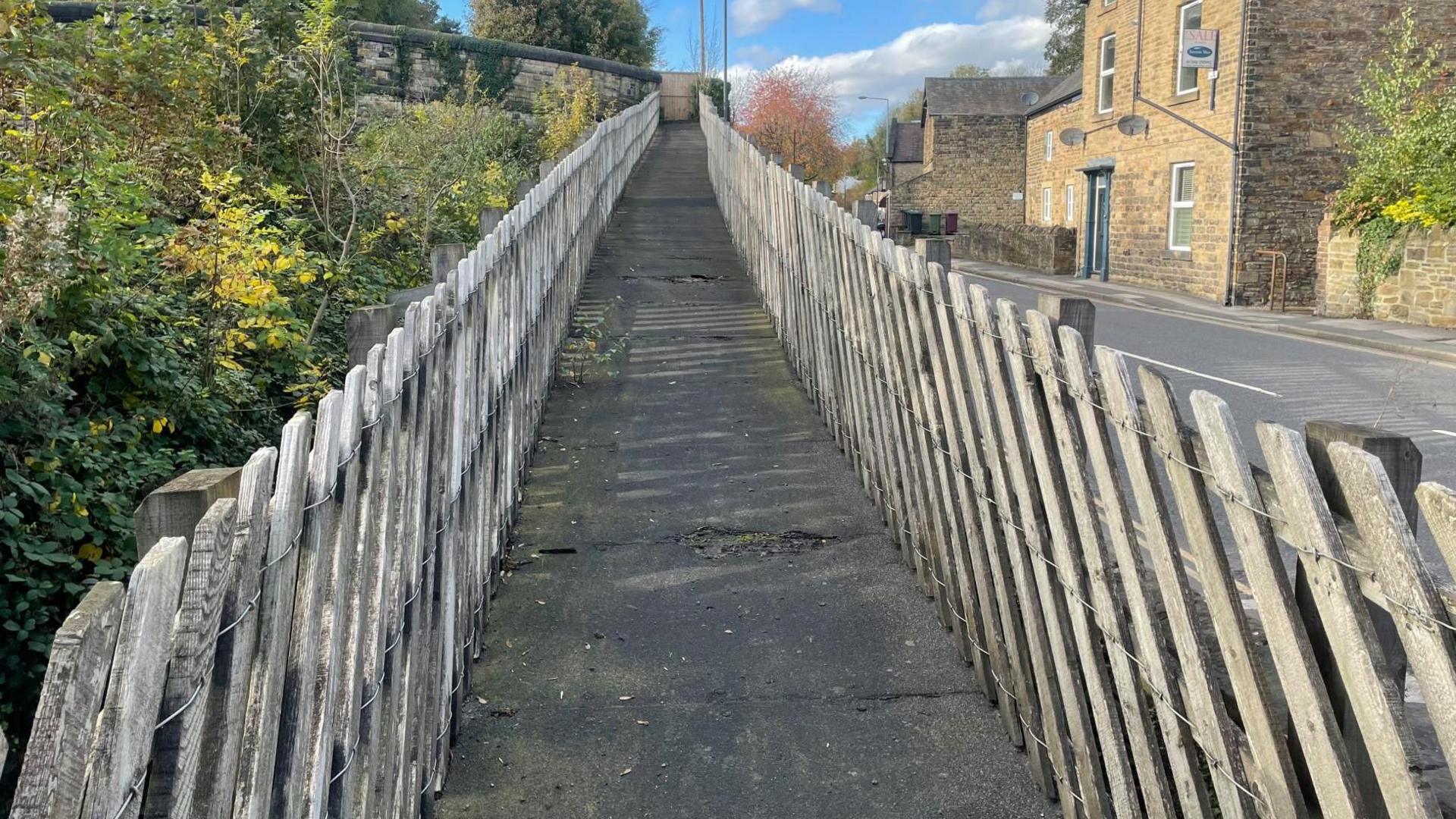 A well worn path leading up to a bridge. Wooden panels on either side.