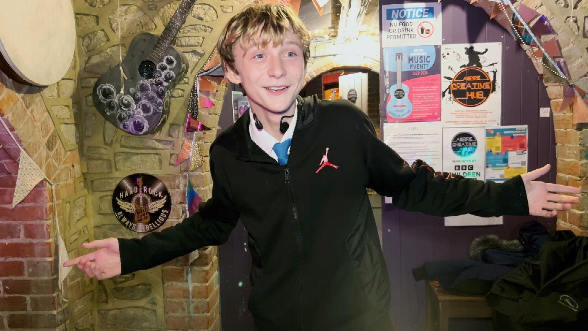 A boy stands in the crypt of an old church where music sessions are help for children. He is standing with his hands stretched out looking happy.