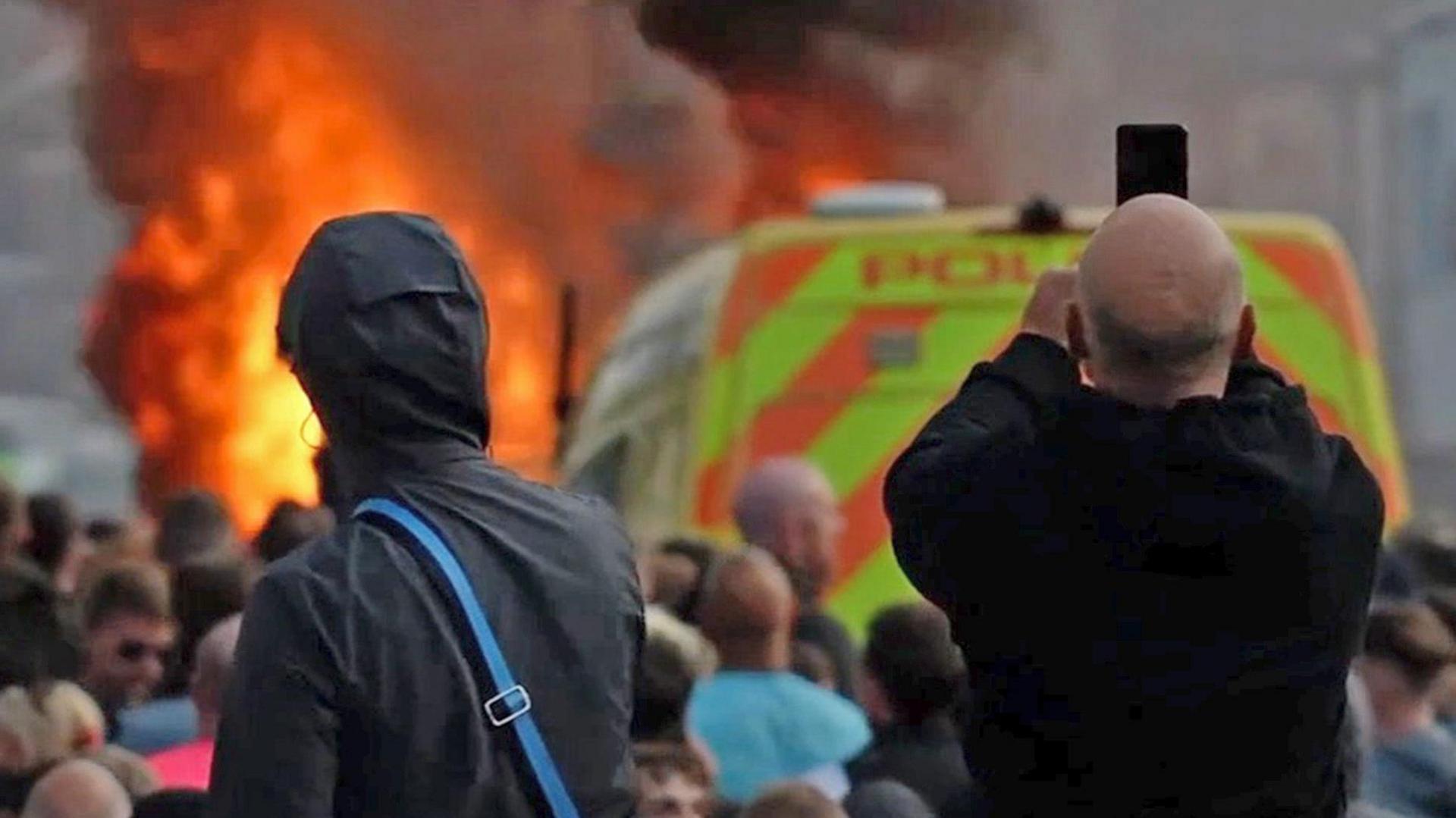 Scene from the riots in Southport in July 2024. Two men are pictured from behind looking towards a police van and flames. One has the hood of his coat up, the other has a bald head and is pointing his mobile phone towards the scene. Between the two men and the fire are a crowd of people. 