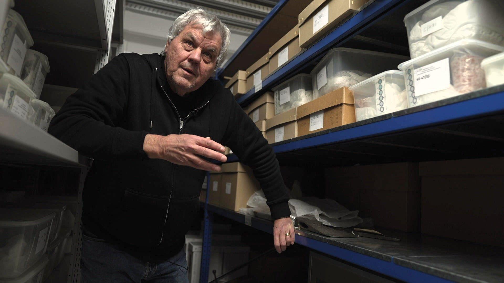 Dyn a gwallt llwyd a dillad tywyll yn sefyll rhwng silffoedd adran archif Amgueddfa Cymru.