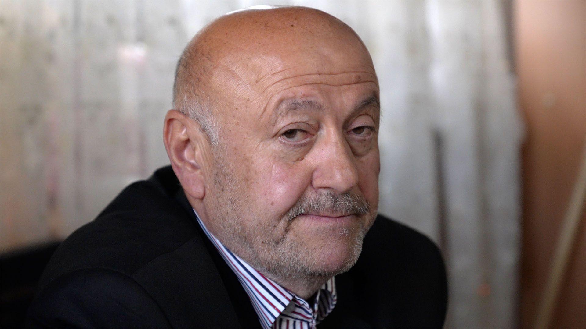 Headshot of a man wearing a white shirt with red and blue stripes with a smart dark jacket. He has a greying moustache, short facial hair and is bald.