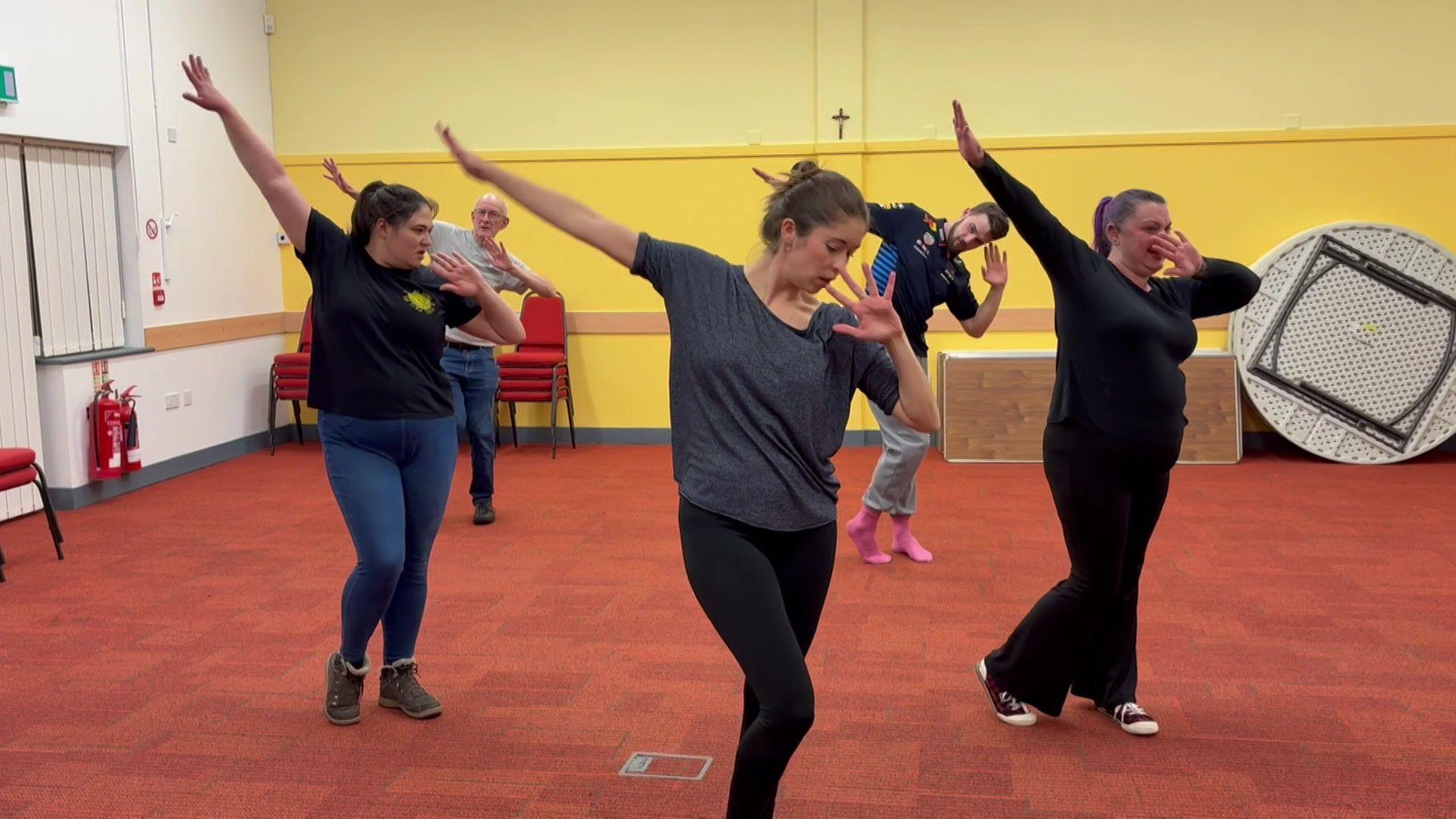 Five people in ordinary clothes dancing in a red-carpeted rehearsal room with yellow and white walls. They are stepping from side to side while holding their right arm up. Their left arms are bent at the elbow and their hands are touching their noses.