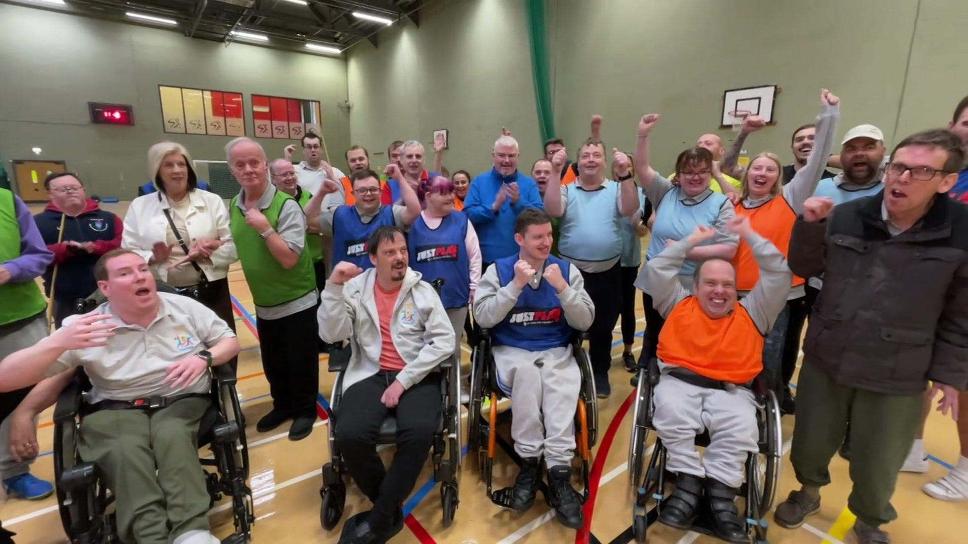 Group of people inside a sports hall cheering. Four people in wheelchairs wearing sports clothes are in the front row with a group of people wearing sports clothes standing behind.