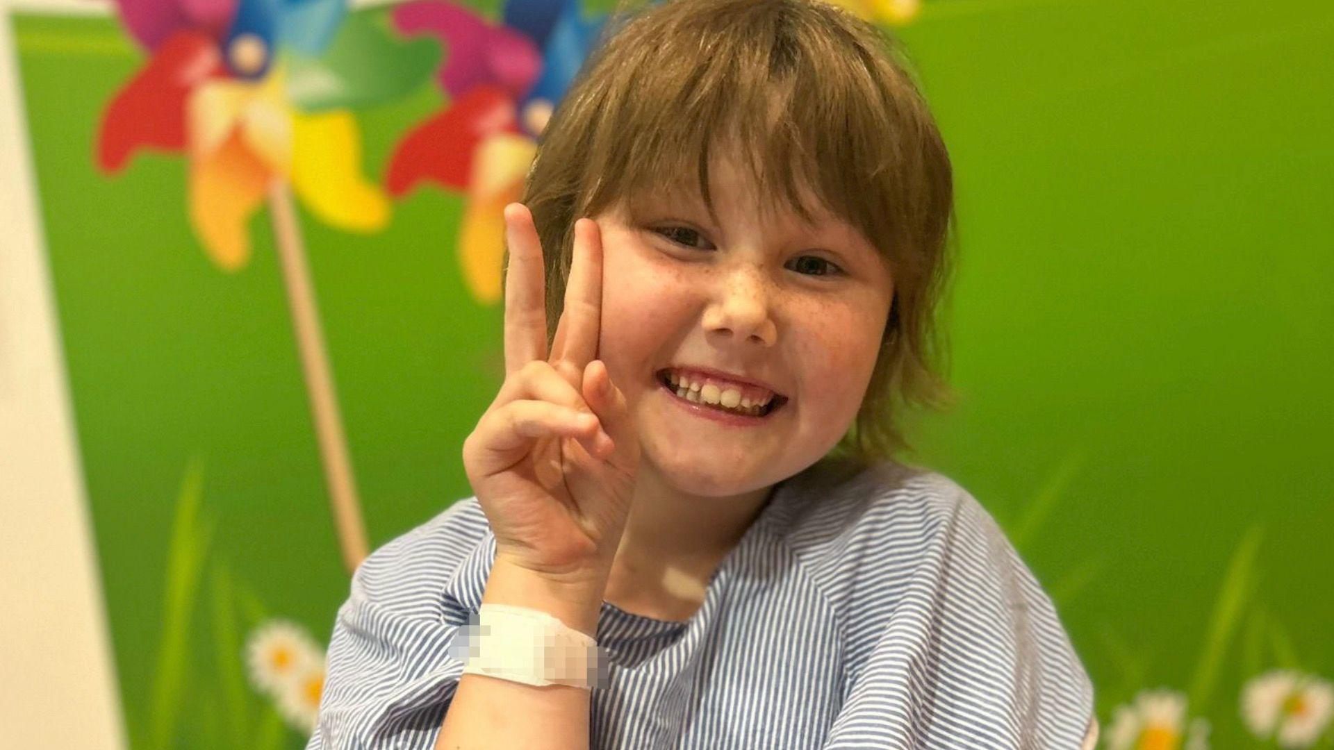 Summer is looking directly at the camera, smiling, making the peace symbol with her fingers. She wears a blue and white striped hospital gown and stands in front of a green wall background in a hospital.