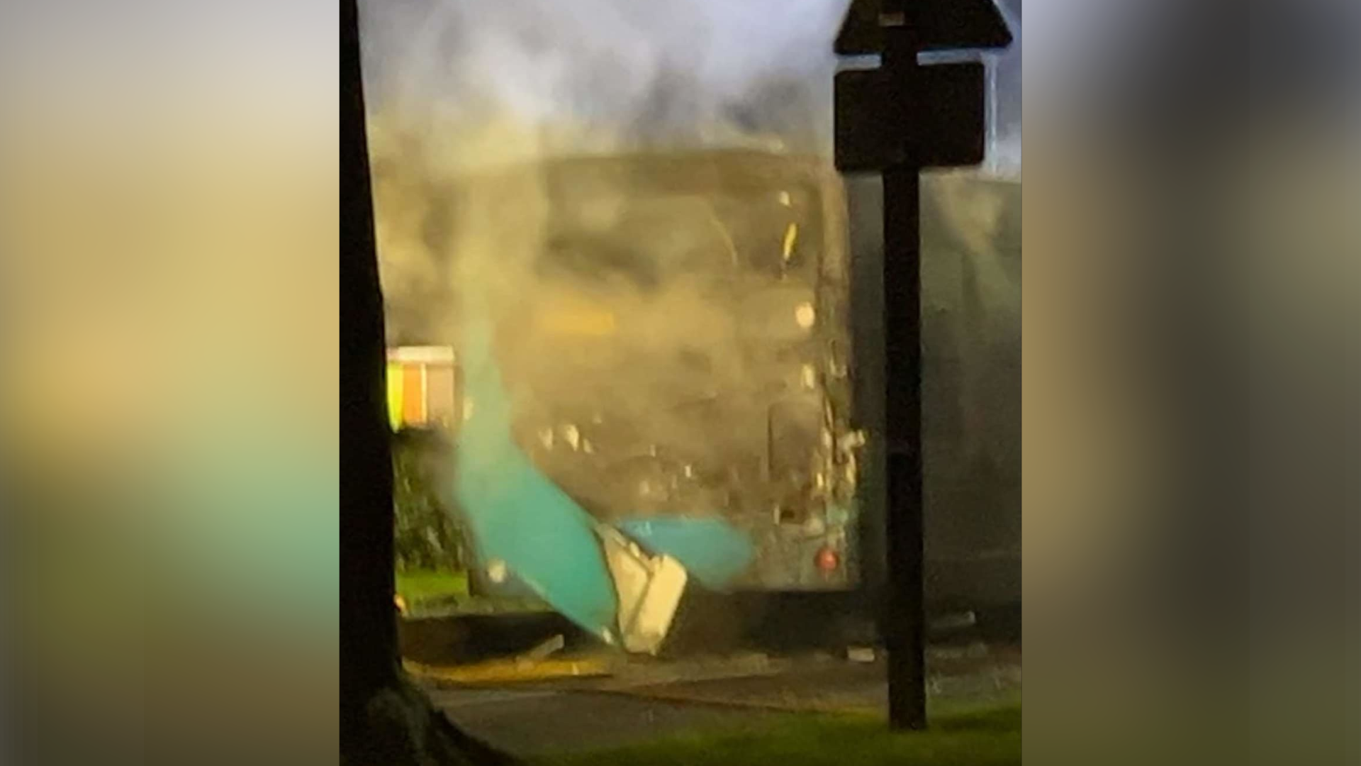 The back end of a bus which is burned out. Smoke is billowing from it and the back pane has been destroyed exposing the inside of the bus. 