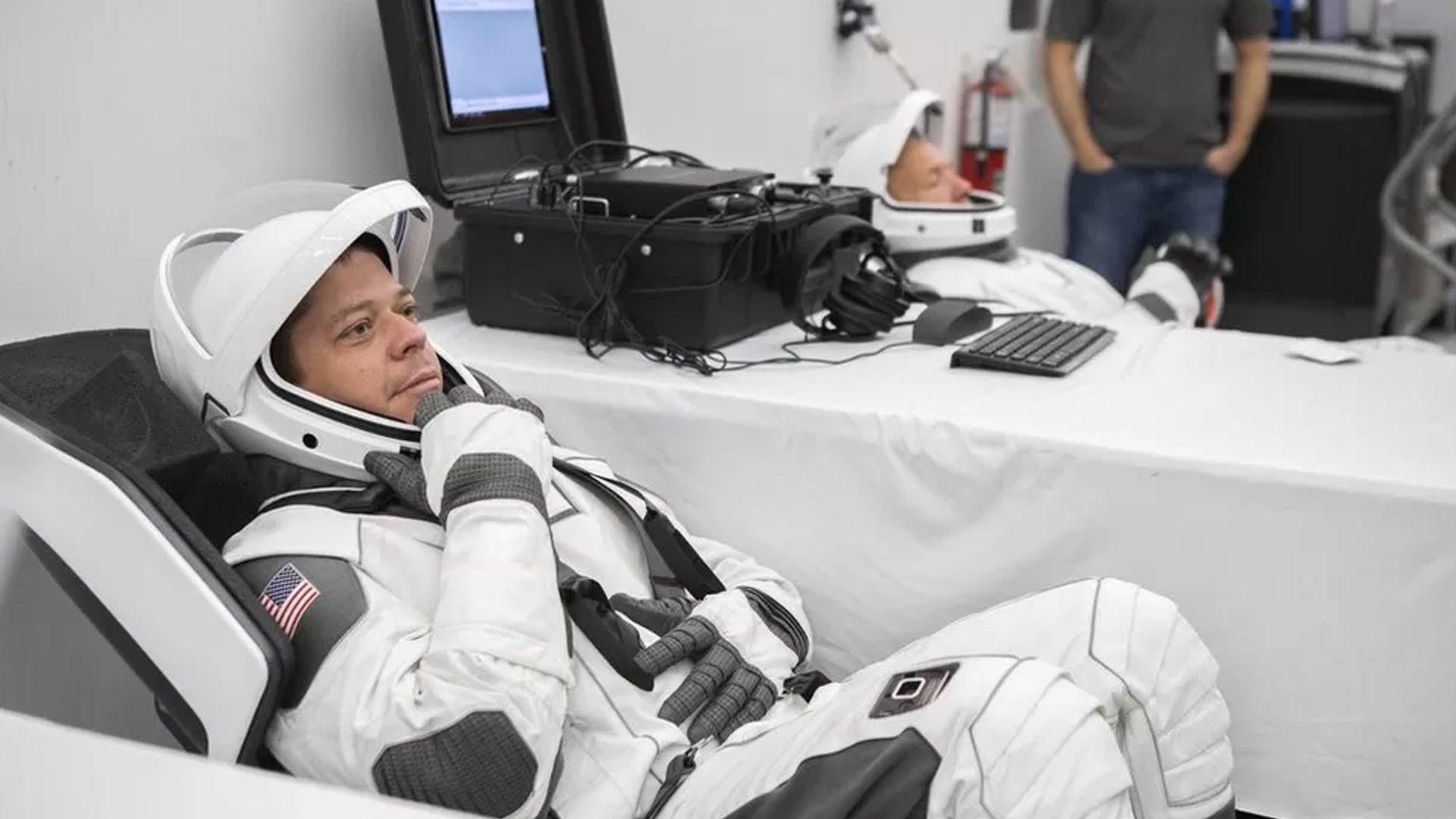 An astronaut wearing a sleek white suit with grey padding sits down inside, with their helmet open