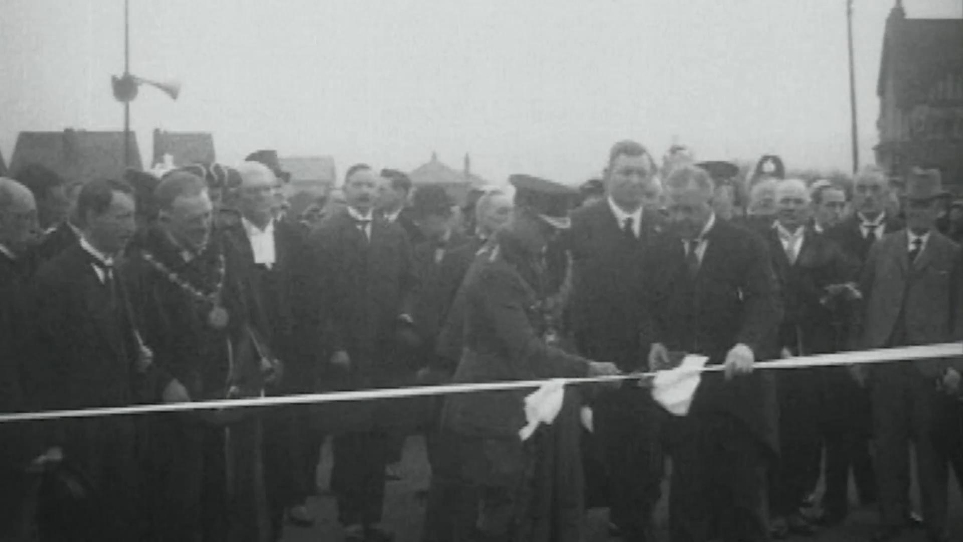 A still from a black and white newsreel film of the Prince of Wales in military uniform cutting a ribbon, accompanied by other men in formal dress, uniform and mayoral regalia.  