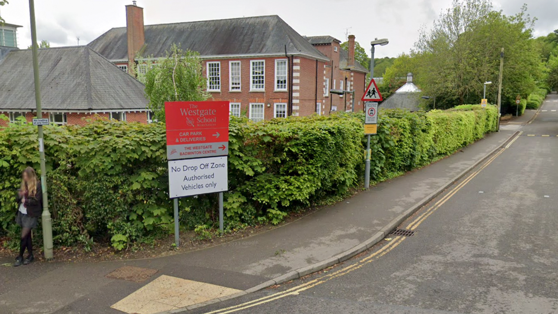 A red school sign saying 'The Westgate School'. The sign is next to a hedge and on the corner of a road. There is a brick building behind the hedge.