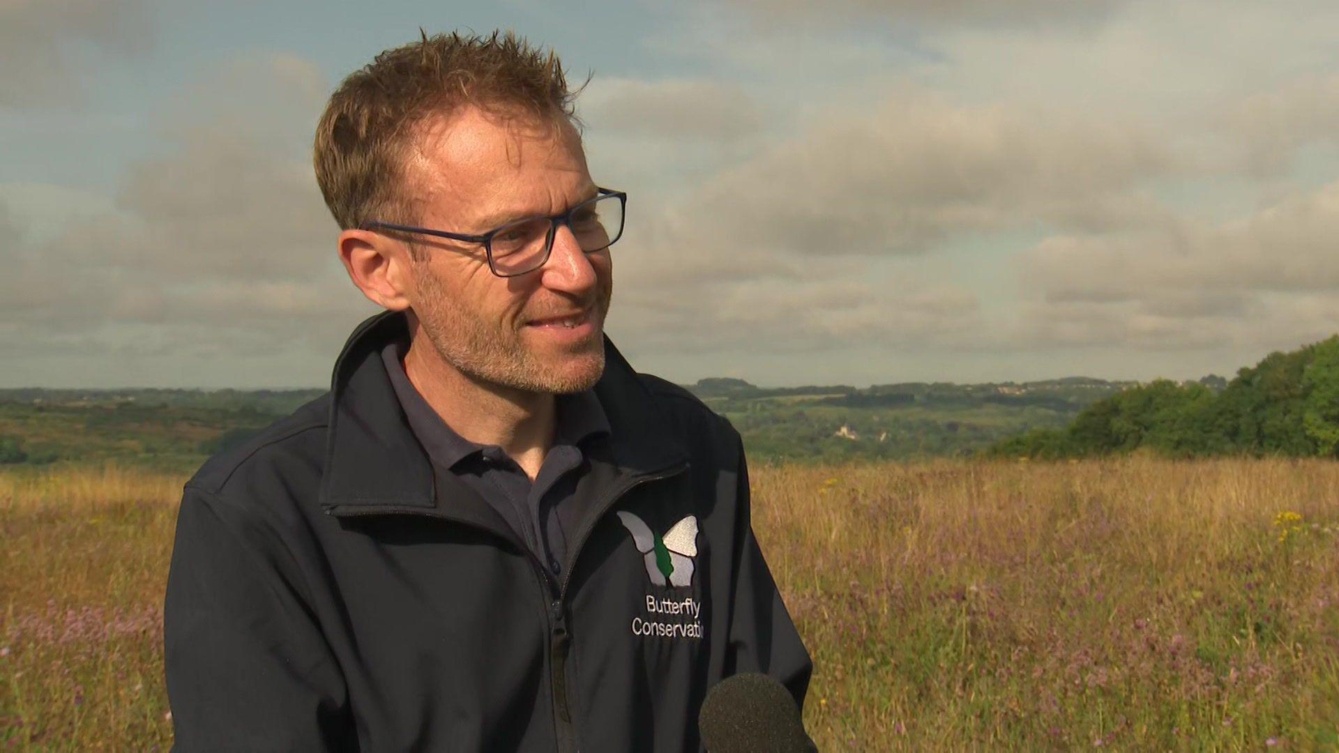 Dr Dan Hoare has short blonde hair and is wearing glasses and a blue fleece. He's standing in a meadow of wildflowers with hills in the distance.