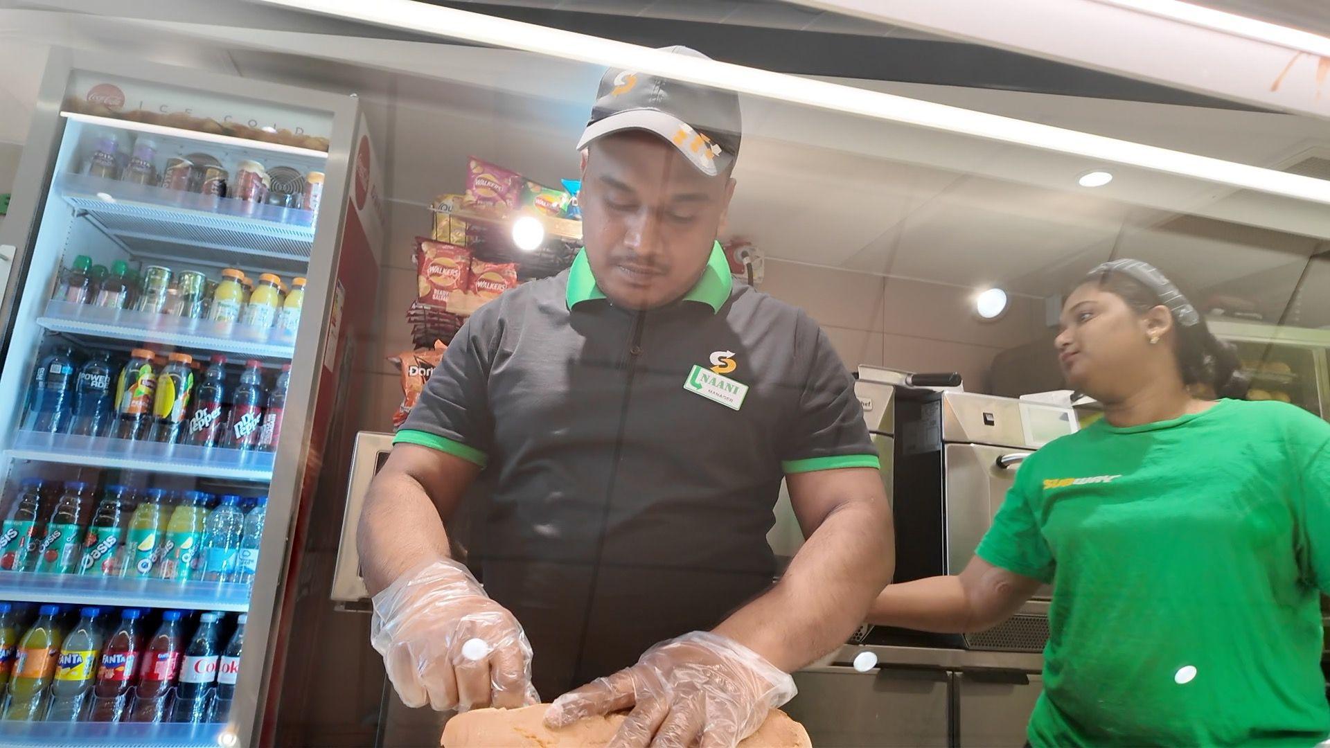 Naani Shaik cutting a subway sandwich behind the counter of his shop. He is wearing a black shirt and black cap. Next to him is a woman in a green tshirt. Behind him is a fridge full of bottles of soft drinks.