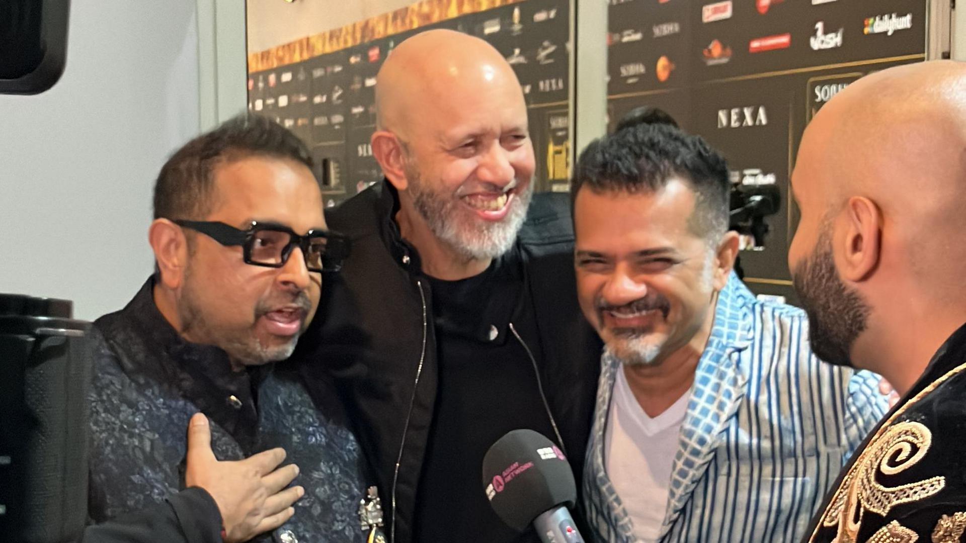 Composing trip Shankar-Ehsaan-Loy smiling as they talk to Haroon Rashid on the IIFA Awards green carpet. Shankar is wearing a dark flower patterned outfit with his arm to his chest, Ehsaan is wearing all black and Loy has a striped blue shirt with a white vest.