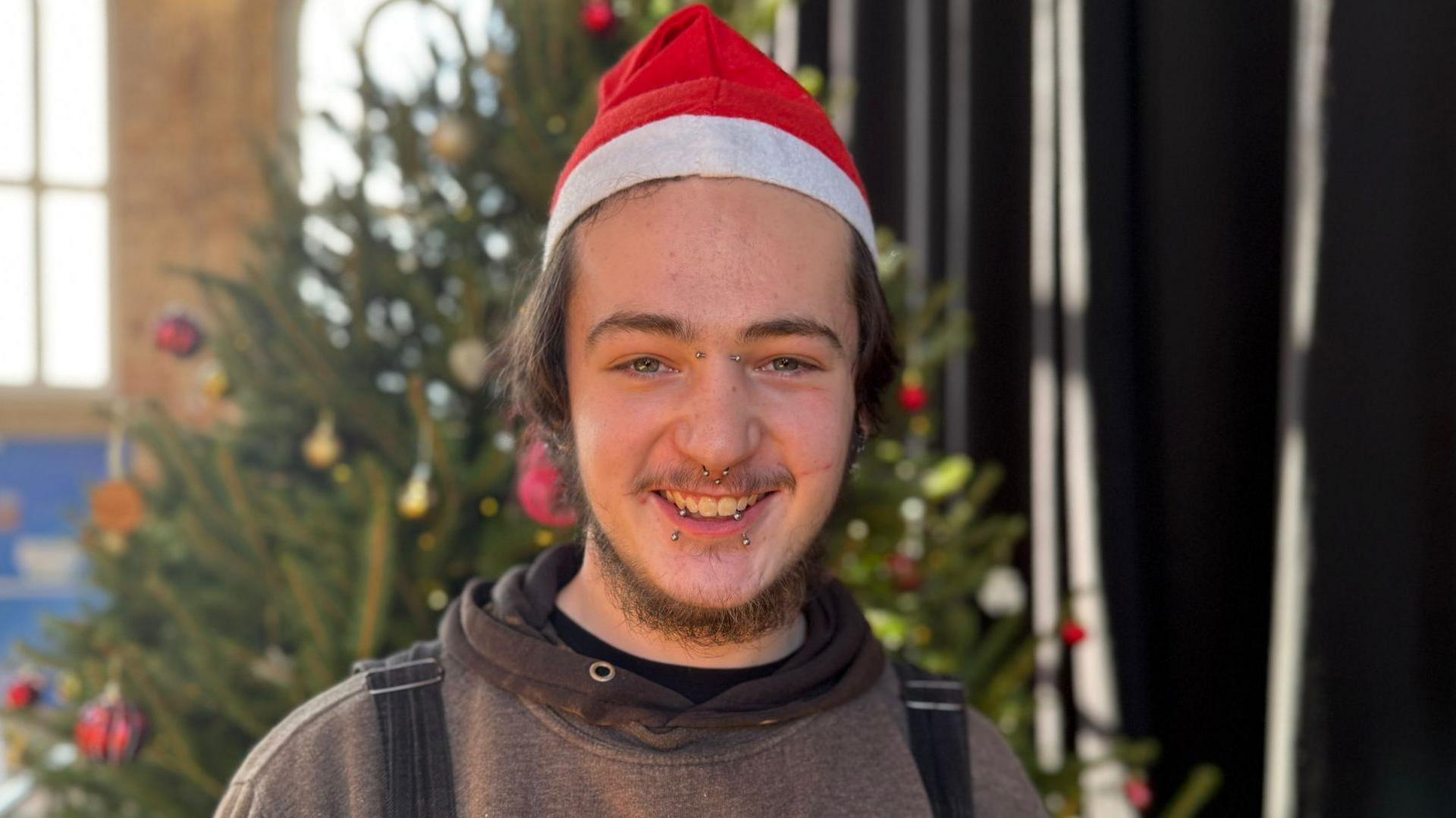 A young man with facial piercings and wearing a santa hat smiles at the camera with a Christmas tree in the background