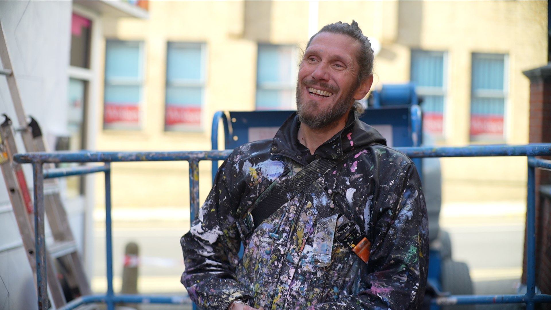 Artist Atma laughs as he looks away from the camera. He stands in front of a cherry picker that rests near the bottom of the building. He wears a black coat that is splattered with different coloured paint. He has long brown hair that has been tied up in a bun on top of his head and he has a thick dark beard.