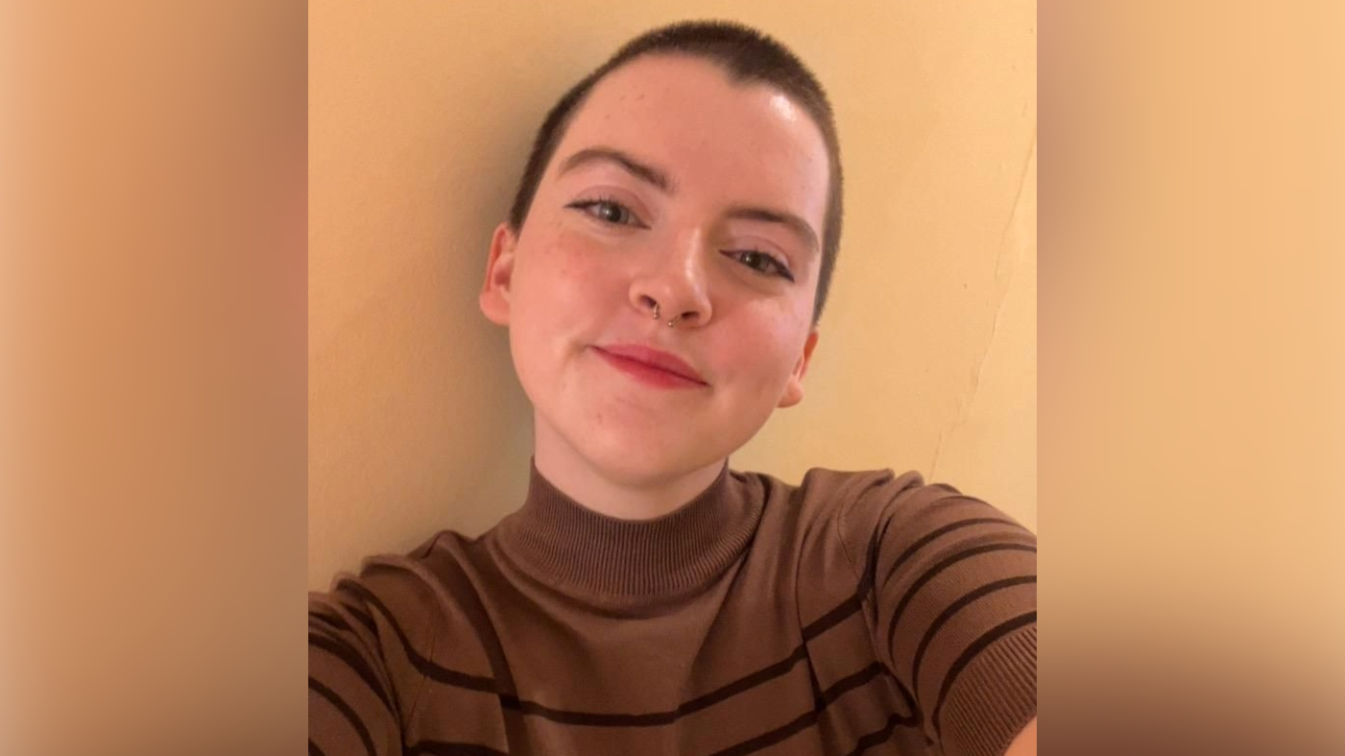 Aoife Kilbane McGowan, 23, is taking a selfie against a beige wall (shoulders-up portrait). She has short brown hair, has a nose piercing and is wearing a brown turtleneck jumper with black horizontal stripes.