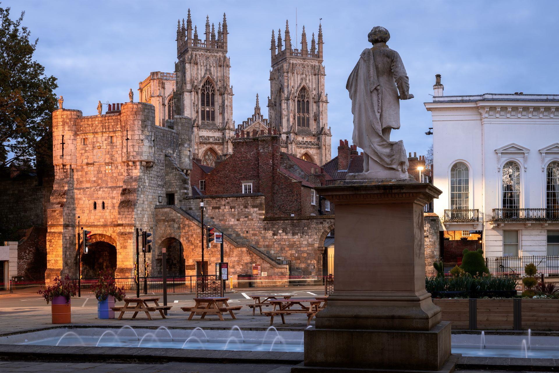 Jets of water fire into a pool at the base of a statue of a man stood on a plinth. In the background the towers of York Minster can be seen.