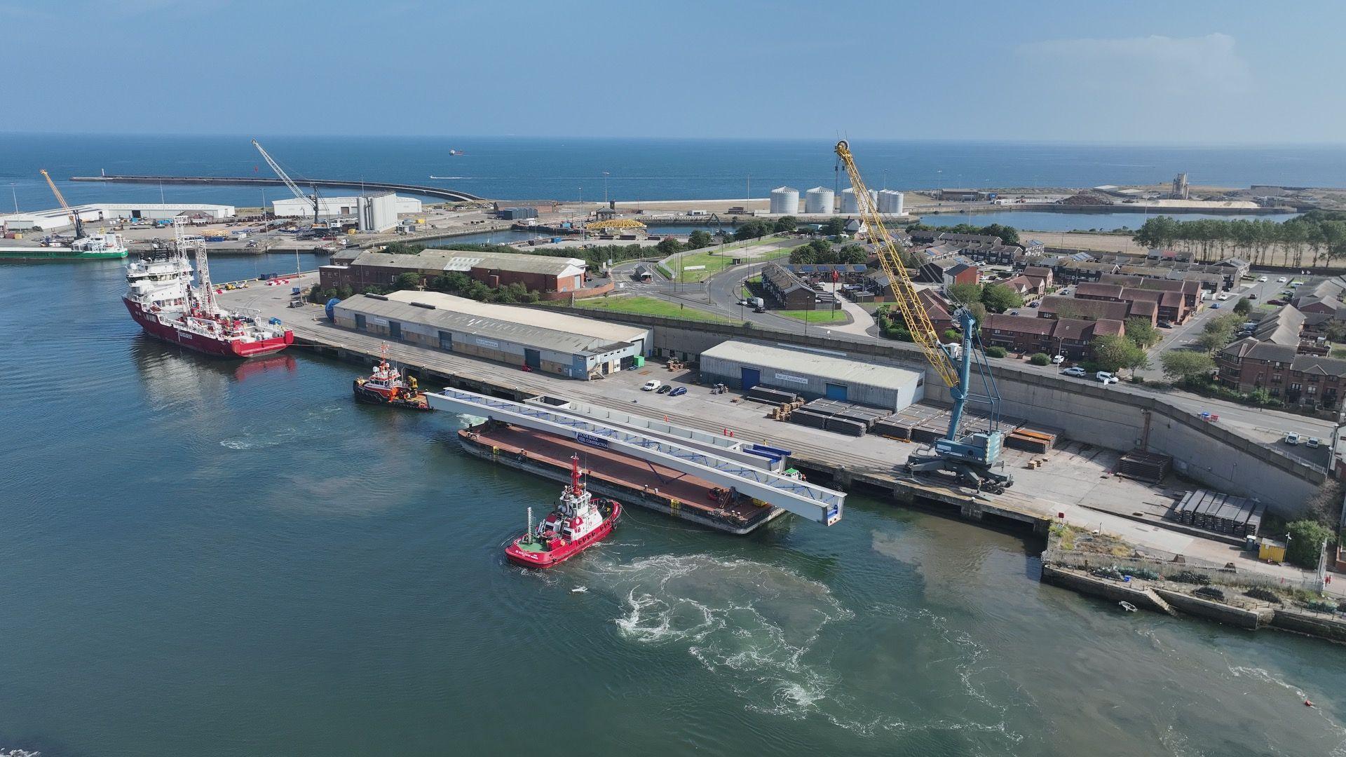 The barge with the steel section moored at an industrial port next to other boats.