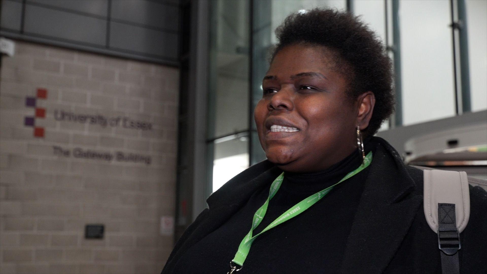 Ojoma Idegwu smiles as she looks to the left of the image in front of the University of Essex Southend campus. She has short black hair and wears a black coat with a black jumper underneath. A green lanyard is around her neck and she carries a grey rucksack on her back. 