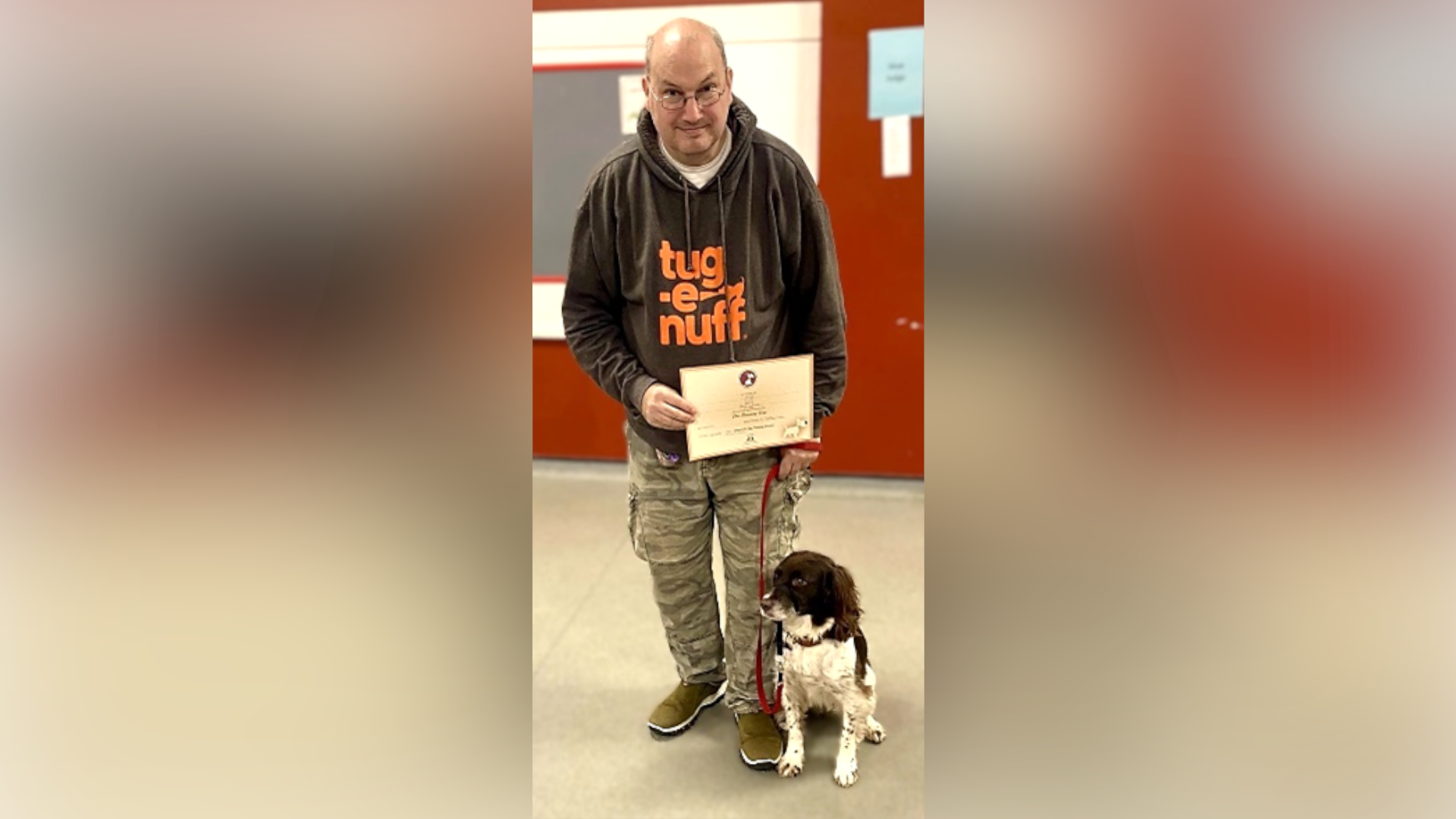 Paul Watts stands with Izzy on a lead by his feat. He holds a certificate. He is bald, wears glasses and a dark grey hoodie with cargo trousers and green trainers.