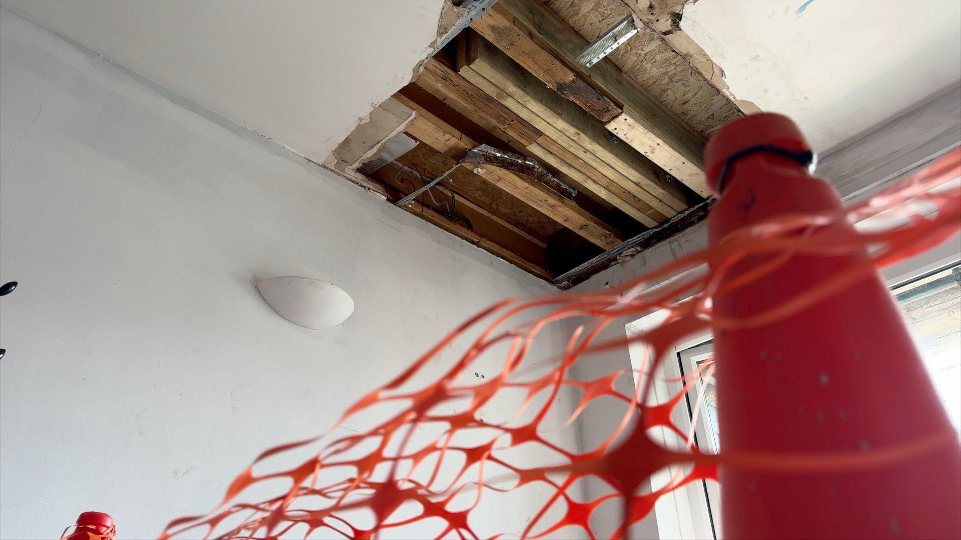 The corner of a room's ceiling with wooden beams and metal structures showing in a large square shape. In the foreground is a warning cone and netting.