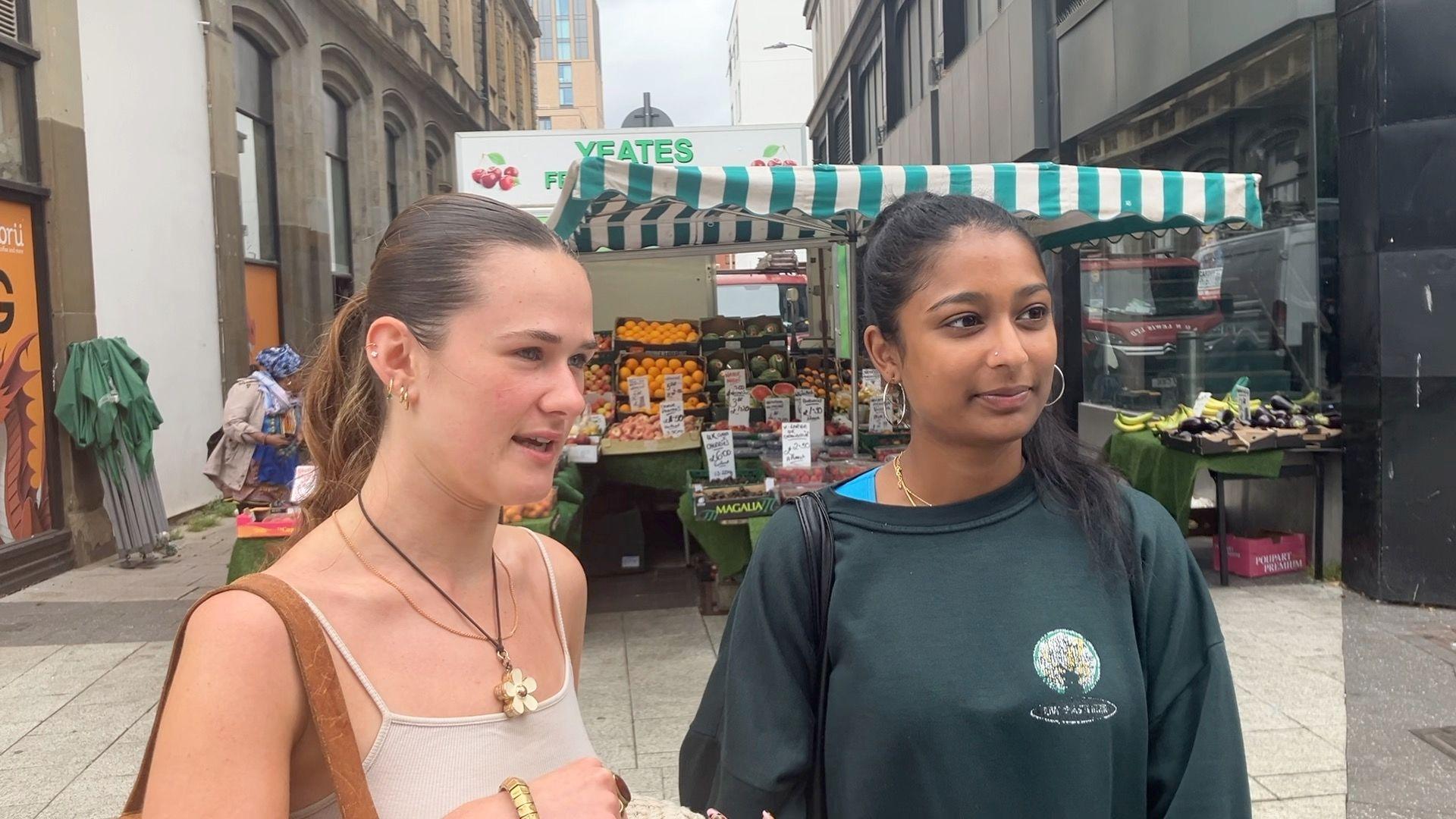 Two girls standing in front of a fruit stall on Cardiff's Queen Street. One is wearing a cream strappy top, large flower pendant and brown shoulder bag, and the other is wearing a dark green sweatshirt, large silver hoop earrings and a black shoulder bag