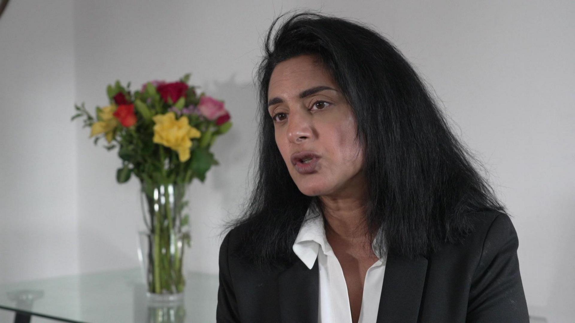 A middle aged woman dressed in a suit jacket and white shirt with long dark hair sits in a neutral setting. There is a vase of flowers on a table behind her.