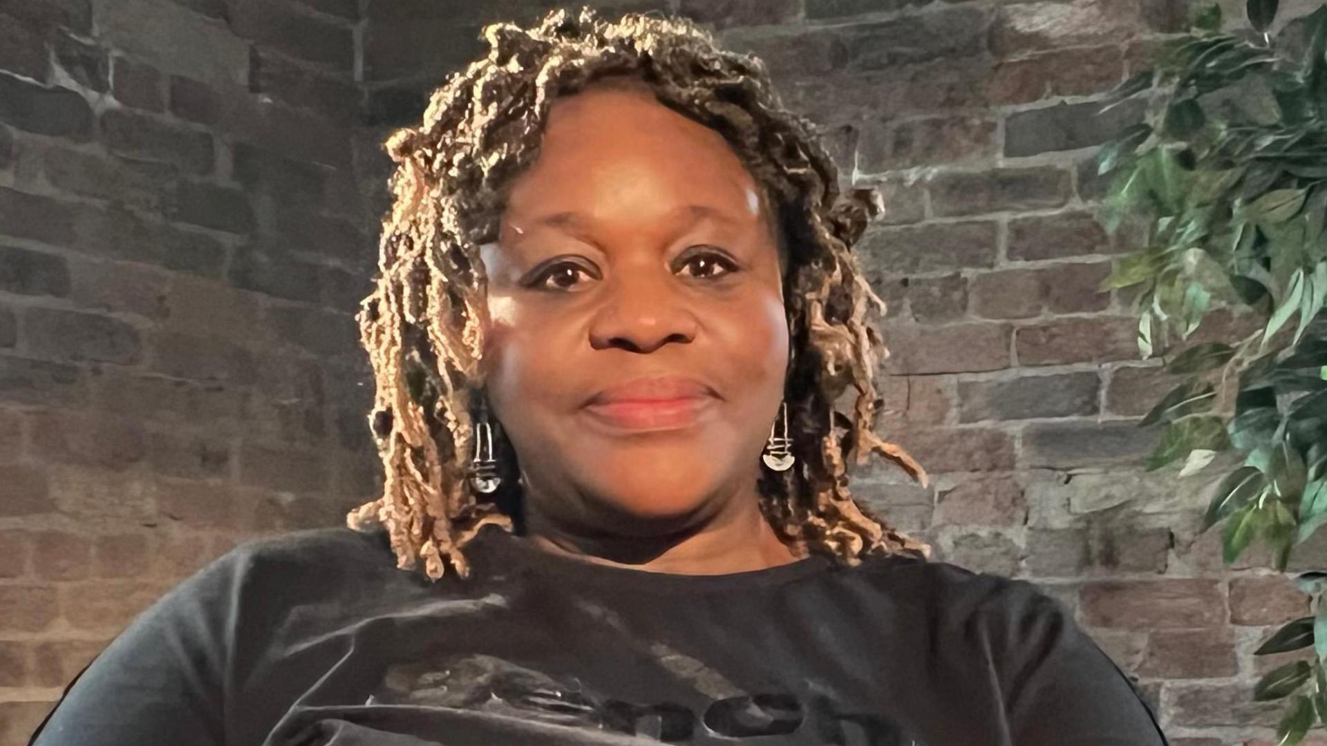 Janet has brown hair, which she has styled in braids. She is wearing a black top and is sitting in front of a bare brick wall. There is a small tree on the right-hand side of her.