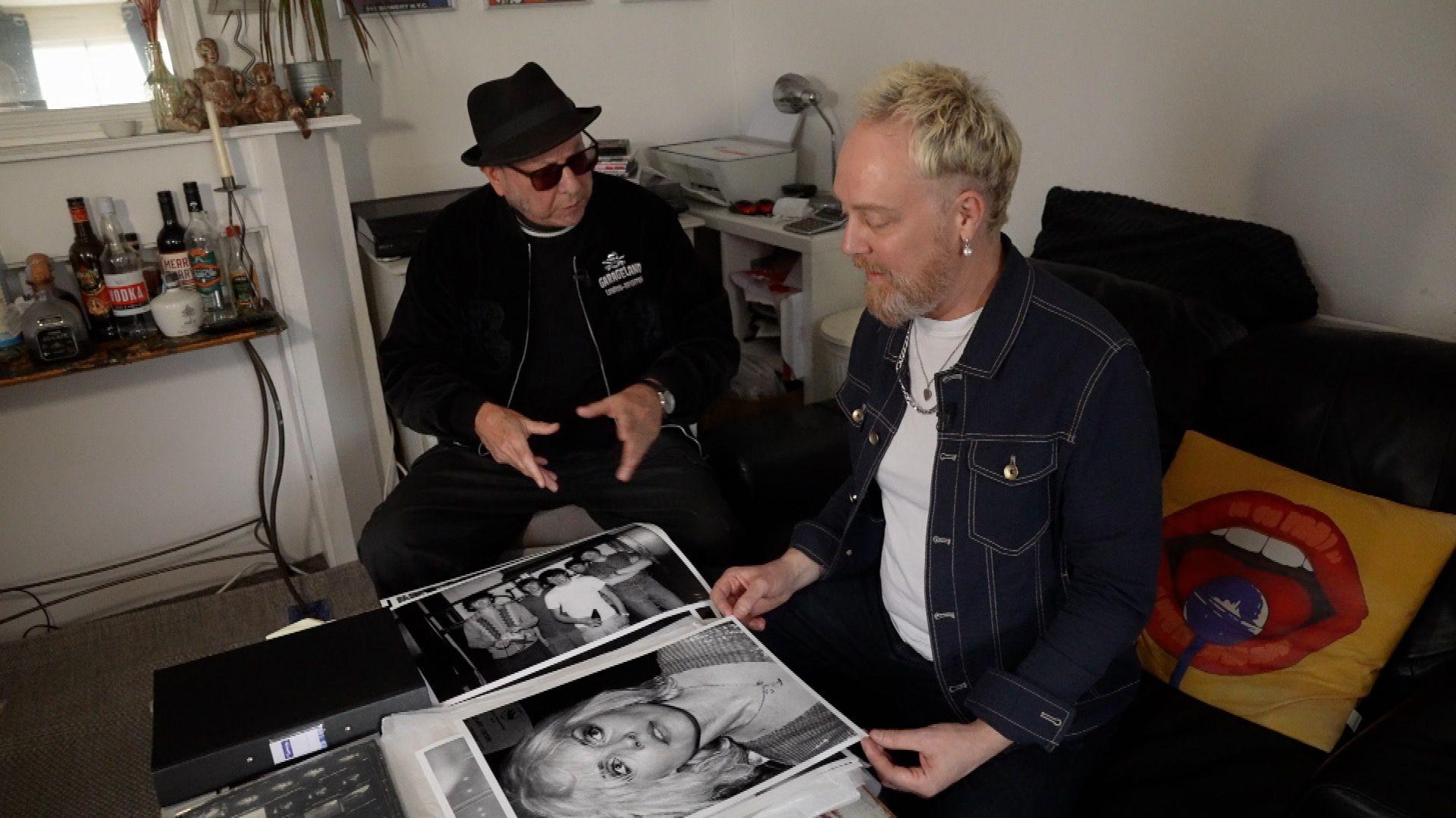 Two men sat on a dark-coloured sofa and talking. One man, in a white t-shirt and denim jacket, is looking through large black and white photographs, which are piled on the table, including of The Undertones and Debbie Harry from Blondie. The other is wearing a black fedora, a black jacket and sunglasses.