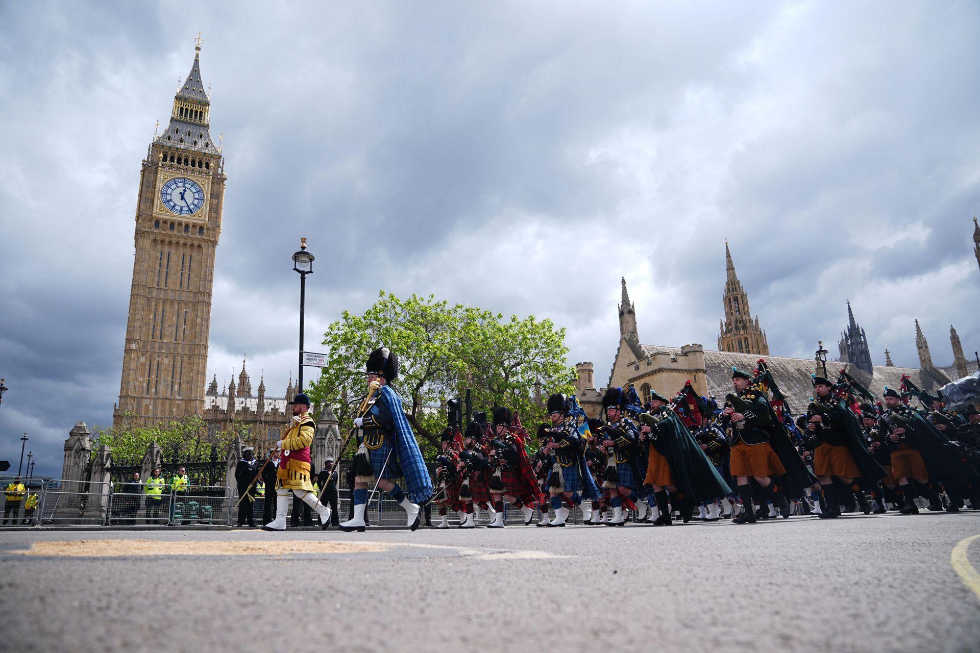 A low-down camera angle shows the military procession marching in Parliament Square outside Big Ben and the Houses of Parliament against a cloudy sky.