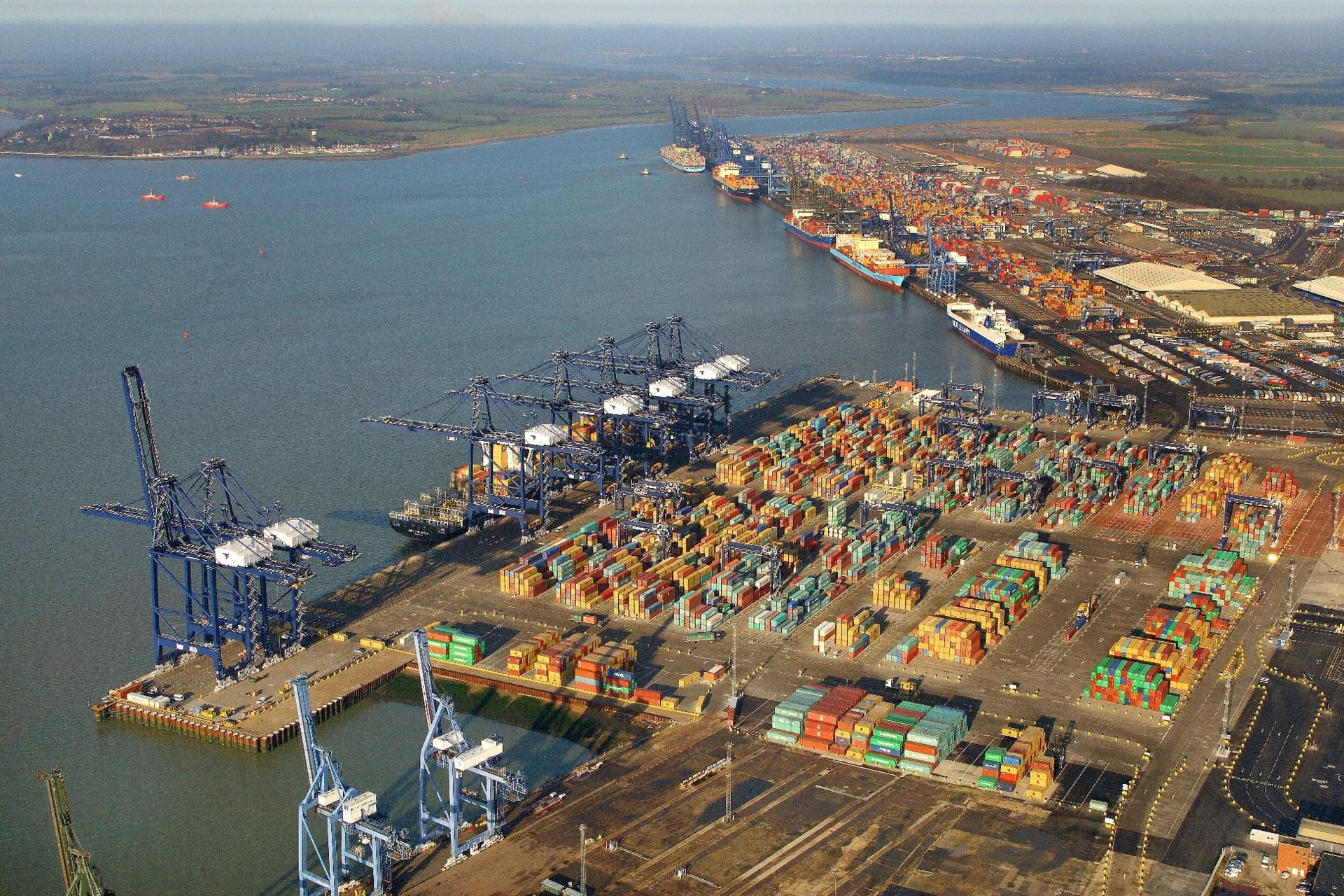 An aerial view of the Port of Felixstowe, showing cranes, containers stacked on the quays and half a dozen ships docked. In the background are is the village of Shotley Gate and the River Orwell.