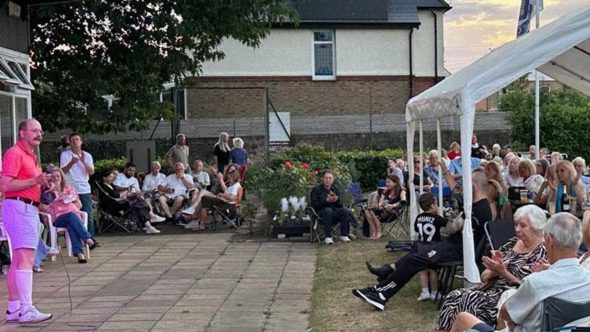A man stood holding a microphone while stood on a patio. He is speaking to a large group of people sat beneath a white gazebo