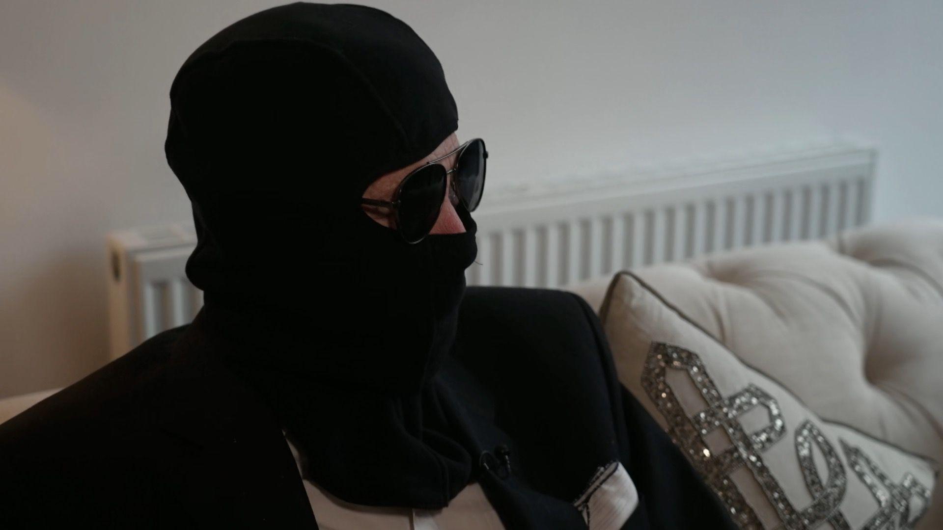 Man sits on a couch while wearing a black suit jacket and a white shirt. To conceal his face, he wears a balaclava, sunglasses and a snood.