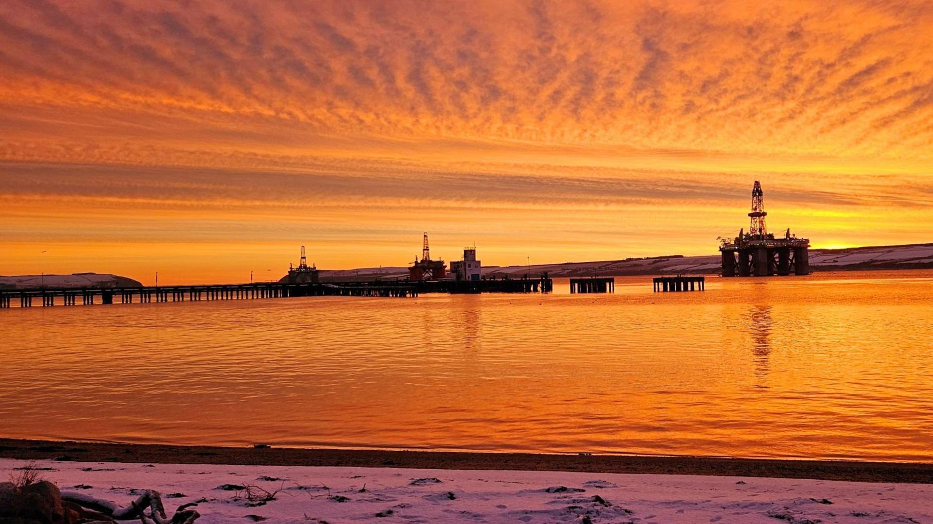 A jetty and oil rigs under the orange glow of the sky. The light is reflected on the waters of the sea. Snow on the shower line has a pink tinge due to the light.