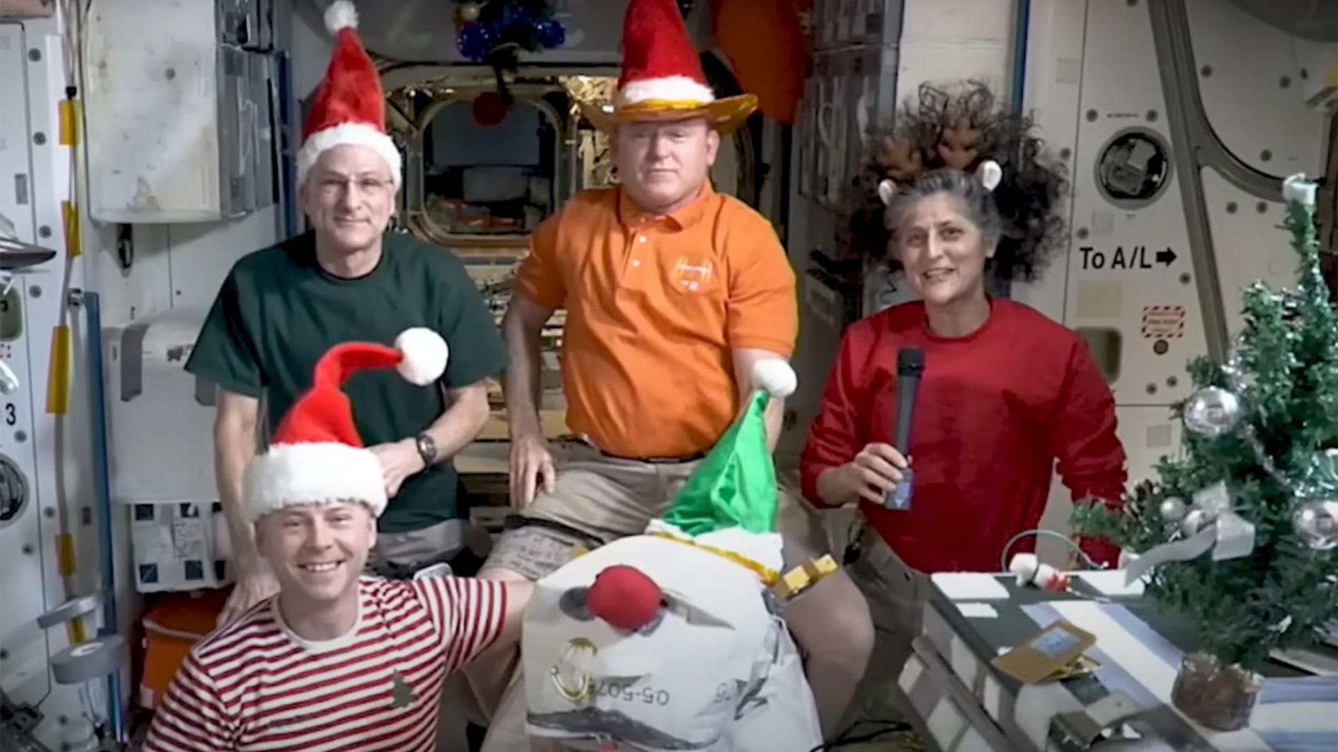 Four astronauts on the International Space Station wearing a mix of Santa hats and reindeer antlers gather together in front of a camera to wish people a merry Christmas