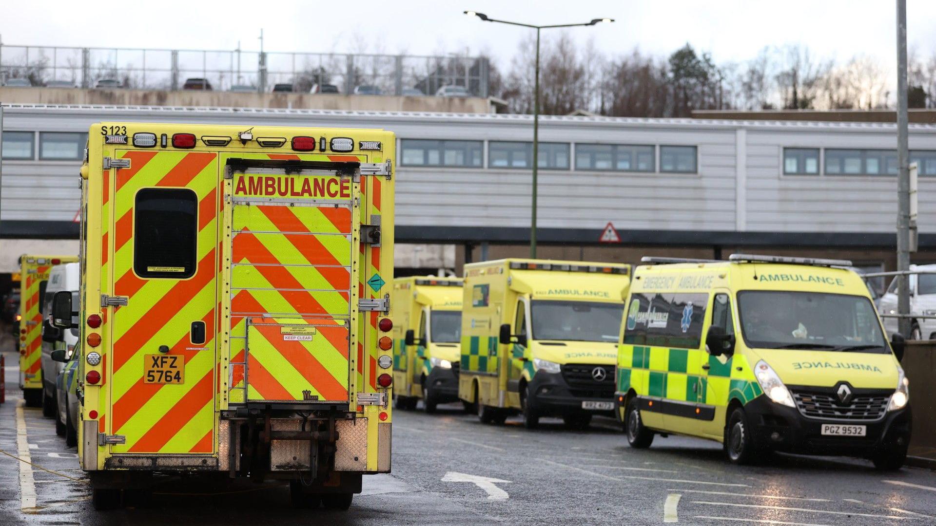A line of yellow and green emergency ambulances is seen parked outside a hospital building. The vehicles are marked with high-visibility red and yellow stickers on the rear, and some display the words "AMBULANCE" on the side. The environment includes a grey, overcast sky, a hospital structure in the background, and street lights. The sky is grey.