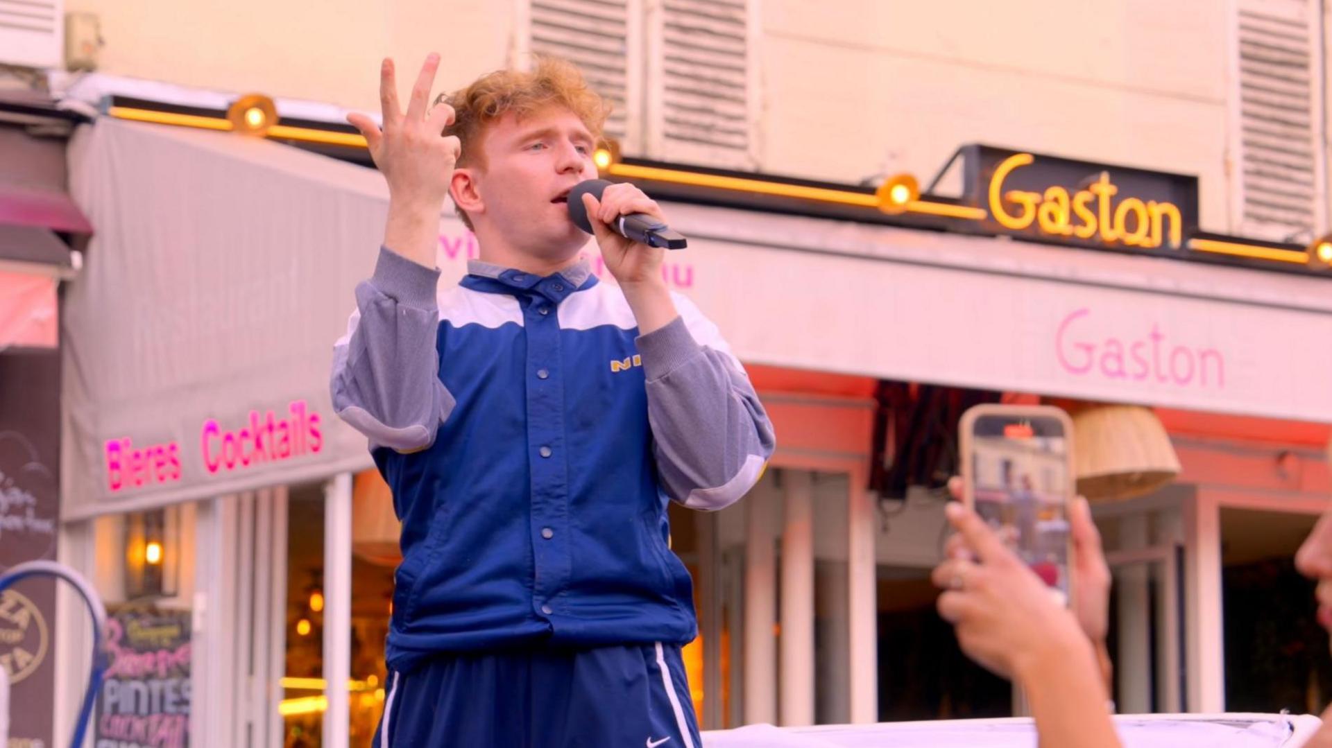 Mickey Callisto dressed in a navy blue track suit belts out Queen's Bohemian Rhapsody in a town square in Paris. 