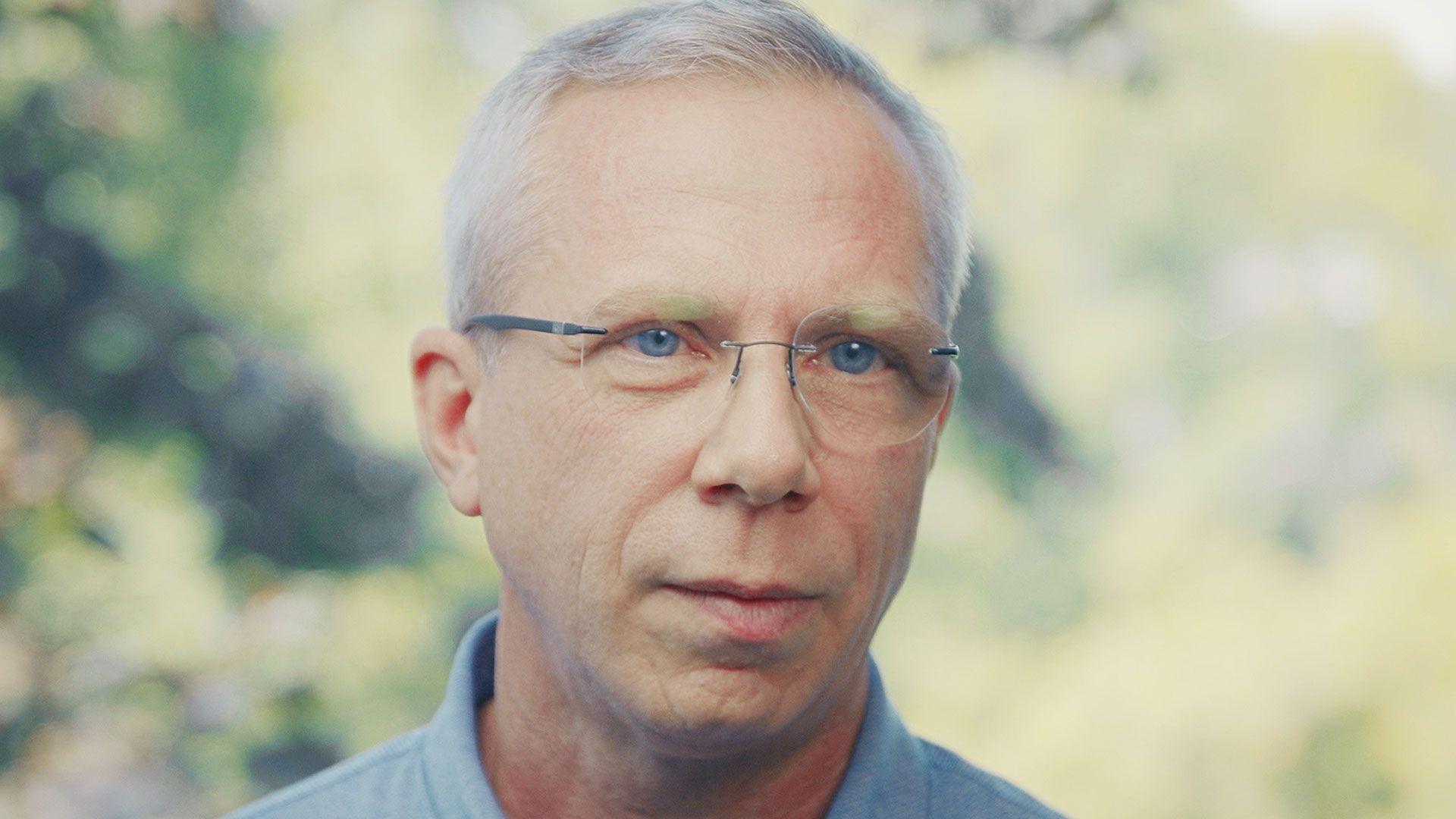 Benoit has rim-less glasses, blue eyes, grey hair and is wearing a blue shirt