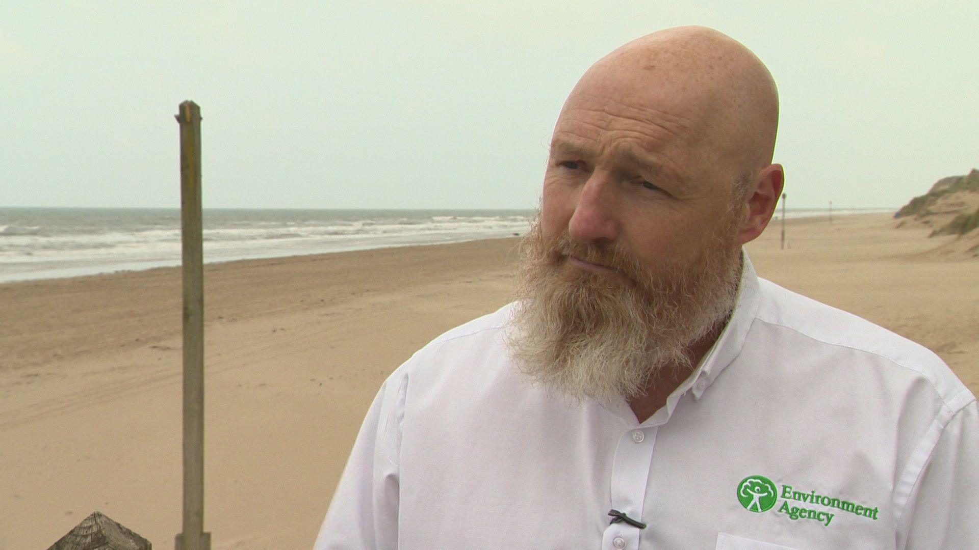 Pete Miles is standing on Formby beach. The sea is to his left and rolling sand dunes are on the right. He is wearing a white shirt with Environment Agency stitched on to the left-hand side in green. 
