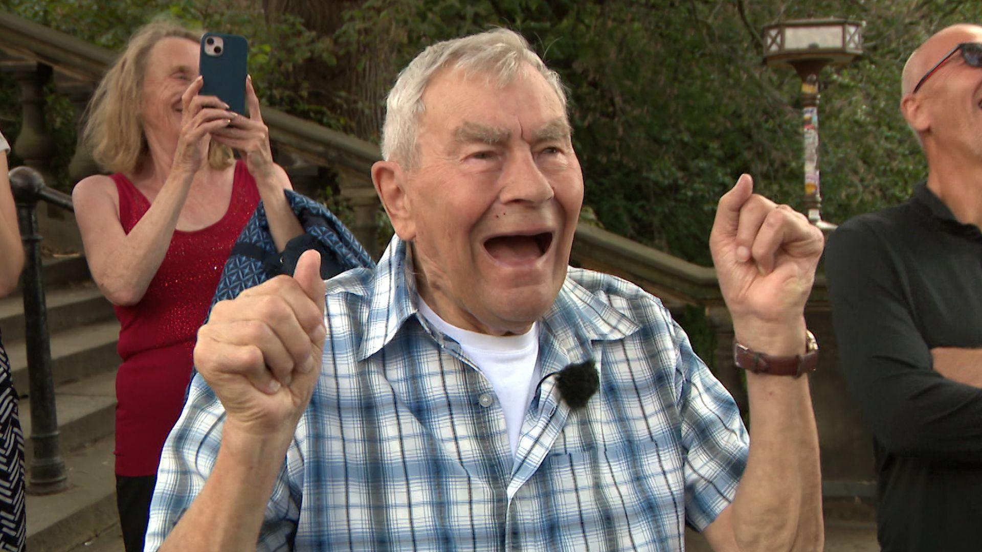 George cheers with his hands lifted as the cuckoo appears. He has grey hair and wears a blue, green and white checked shirt. 