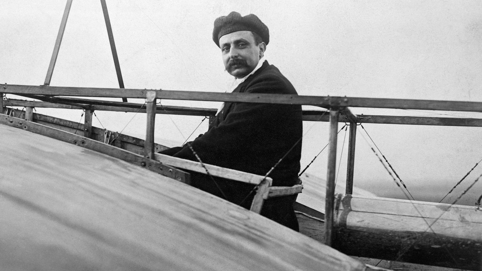 A black and white photograph of Louis Bleriot, wearing a beret and a moustache, sitting in the cockpit of his monoplane.