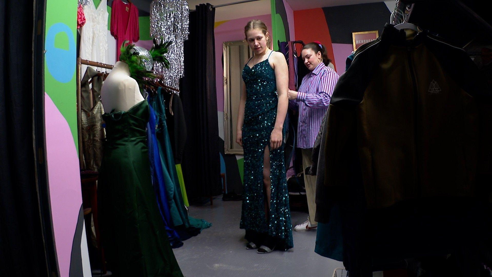 A girl tries on a dress in the shop with the help of a volunteer shop worker. She is wearing a dark green sequin dress. 