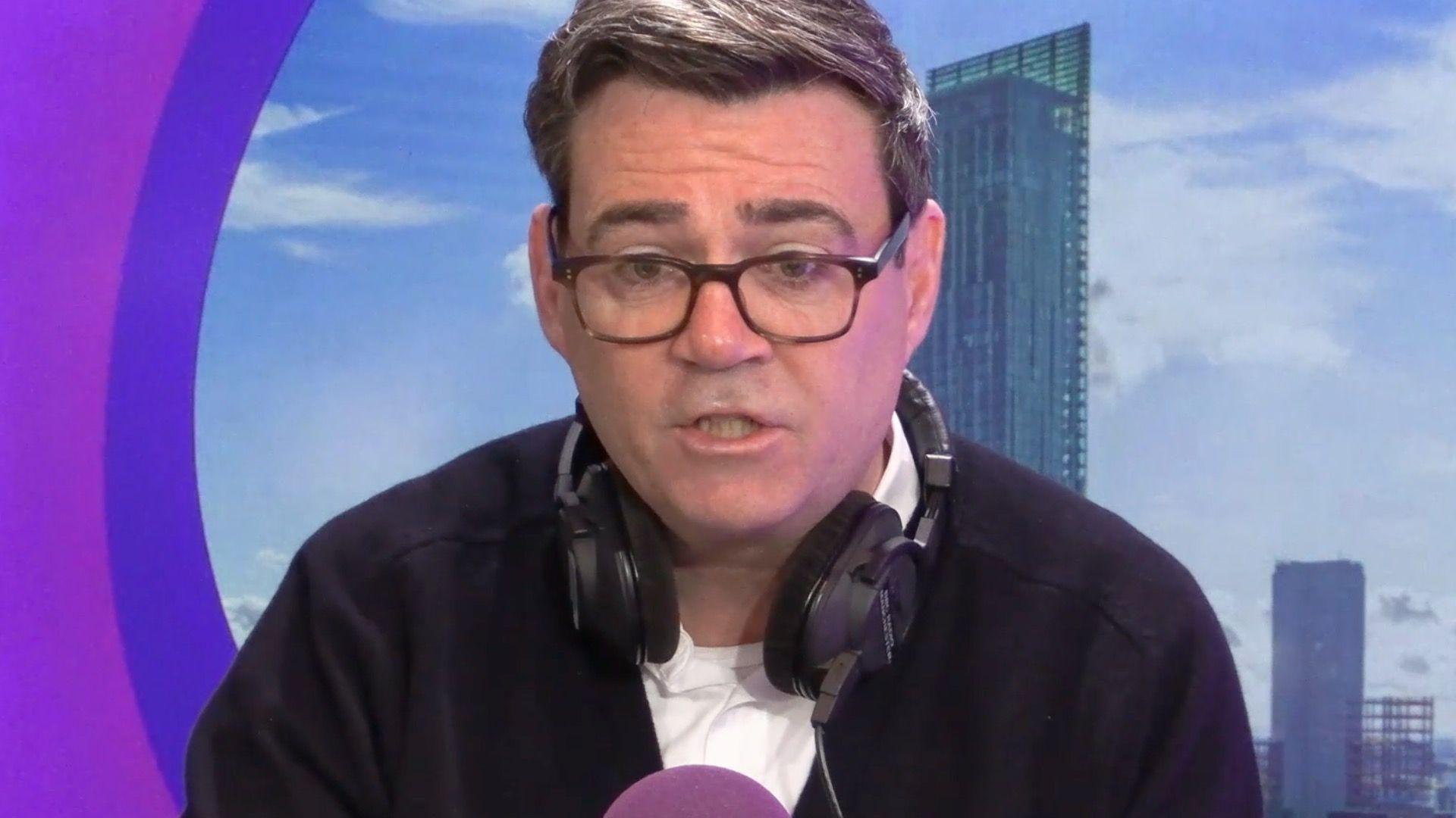 Andy Burnham on radio manchester with purple background. He has dark rimmed glasses a black jacket and white T-shirt